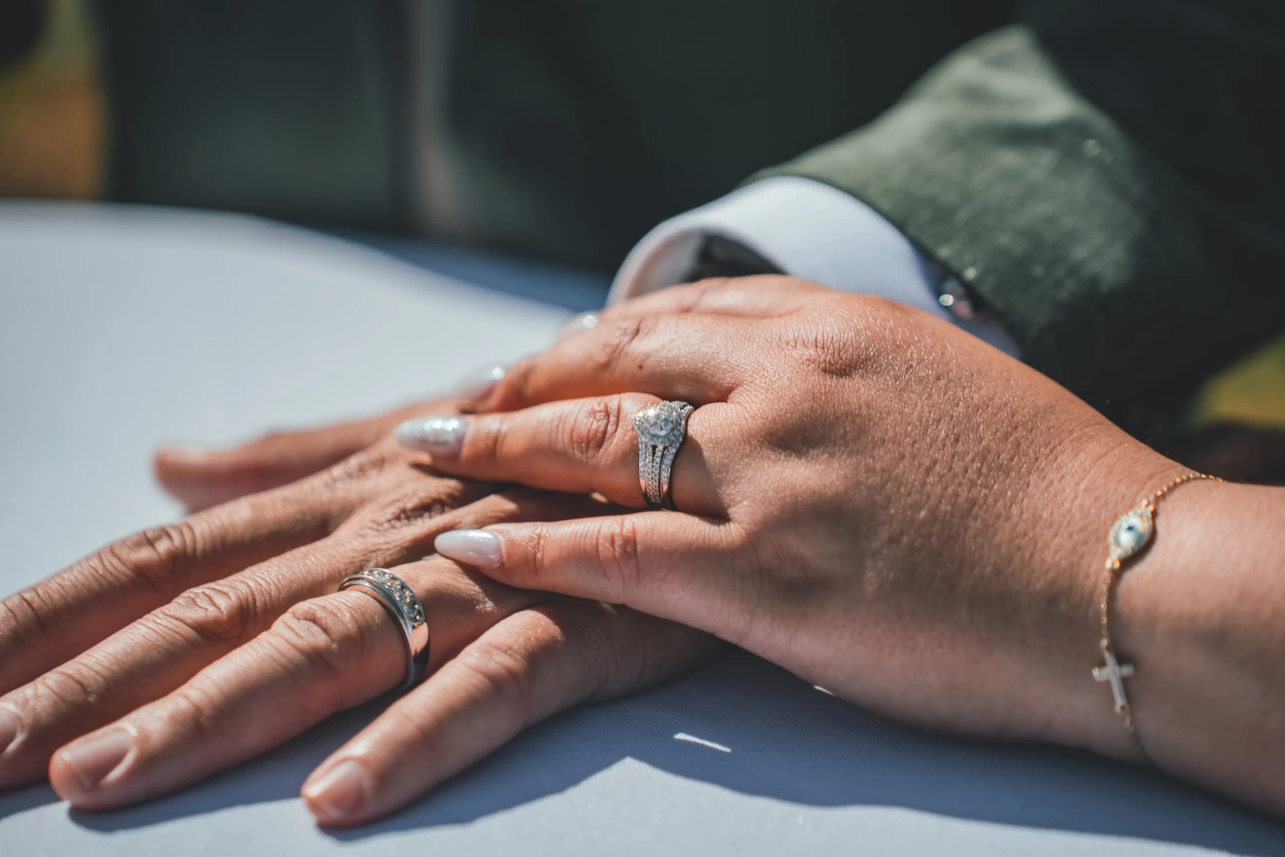 Close-up of two hands with wedding and engagement rings resting on a surface, with a blurred background of someone in formal attire.