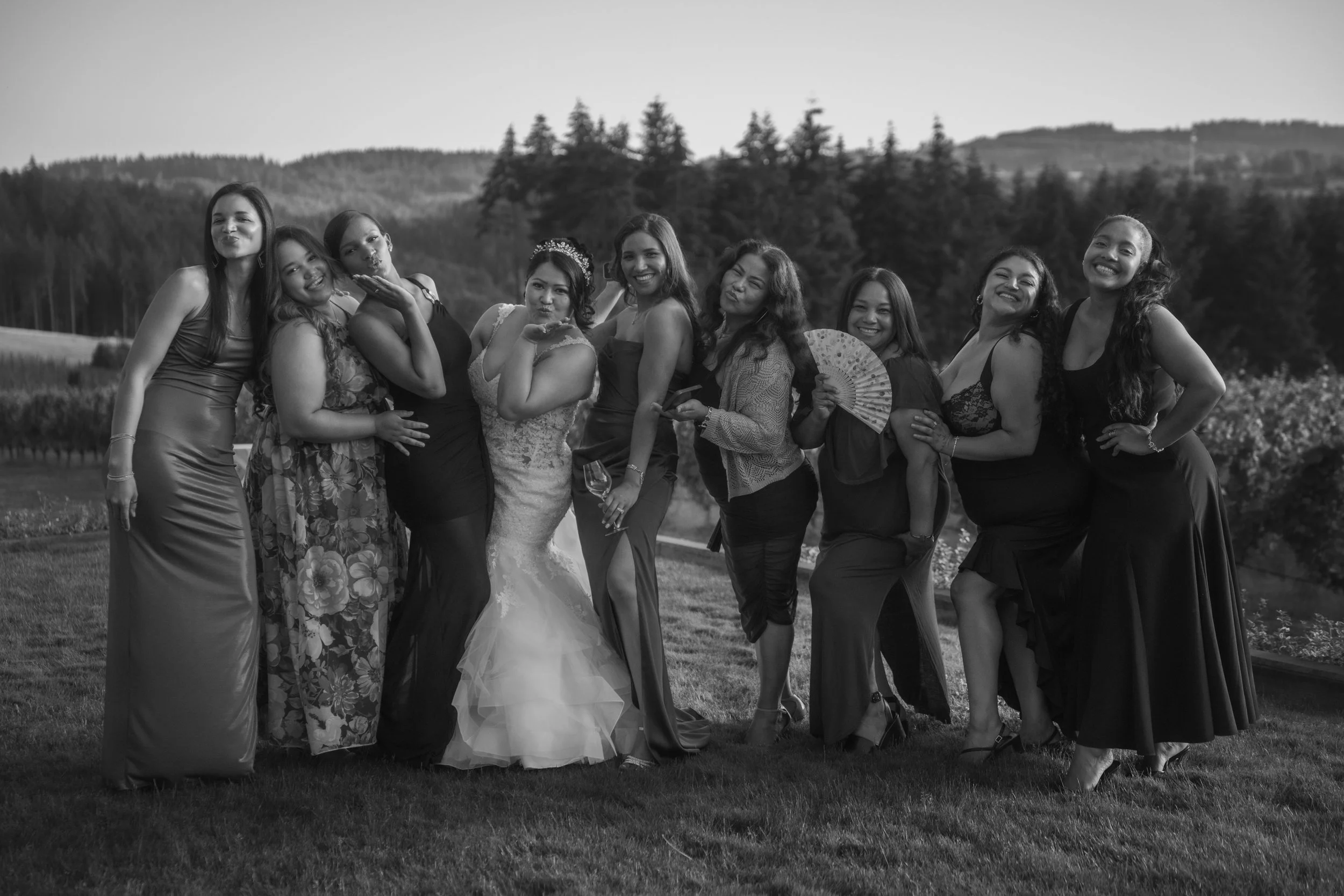 A group of ten women dressed in formal and semi-formal attire, smiling and posing together outdoors in front of a scenic landscape with trees and hills.