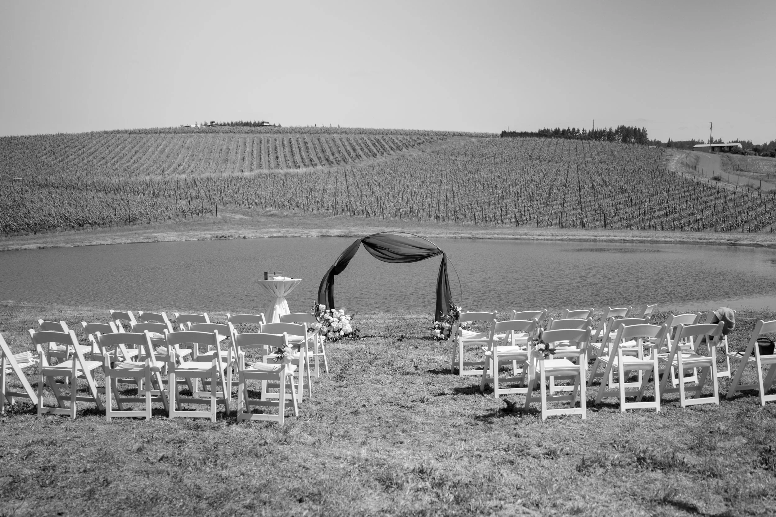 An outdoor wedding setup by a lake with a row of white chairs, an arch draped with cloth and flowers, and a view of rolling vineyard hills in the background.