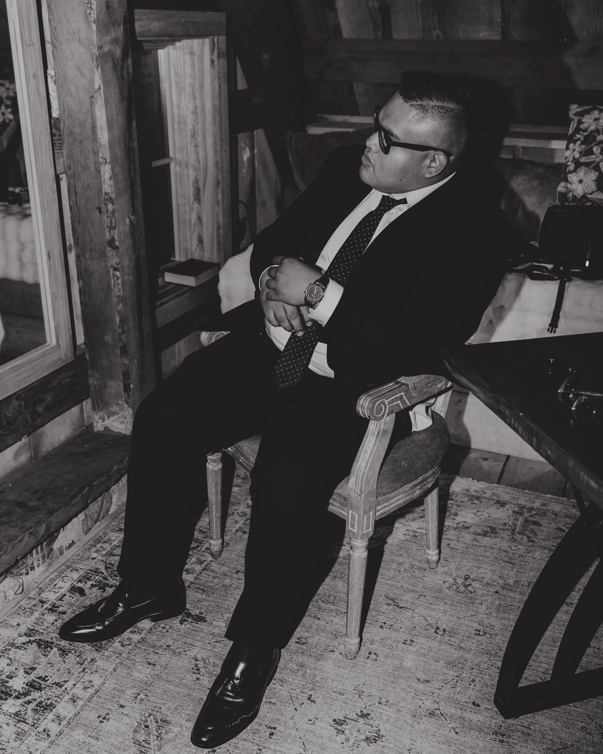 A man dressed in a business suit with a polka dot tie, wearing glasses, sitting on a wooden chair with his hands clasped, in a rustic indoor setting with wooden walls and a wooden table nearby.