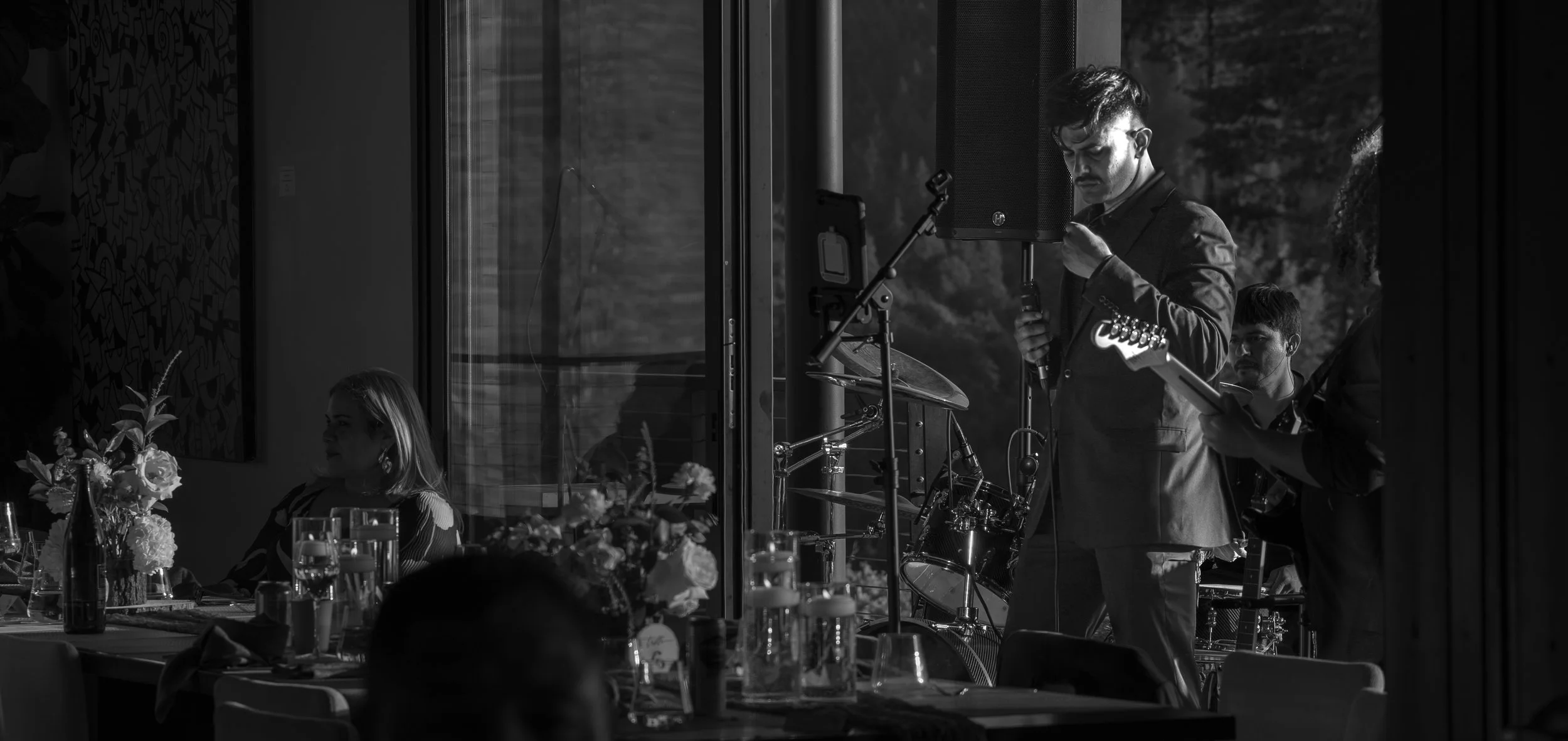 Black and white photo of a musician with glasses and a mustache playing a guitar, standing next to a drummer, while a woman sits at a table with floral centerpieces and glasses in a venue with large windows.