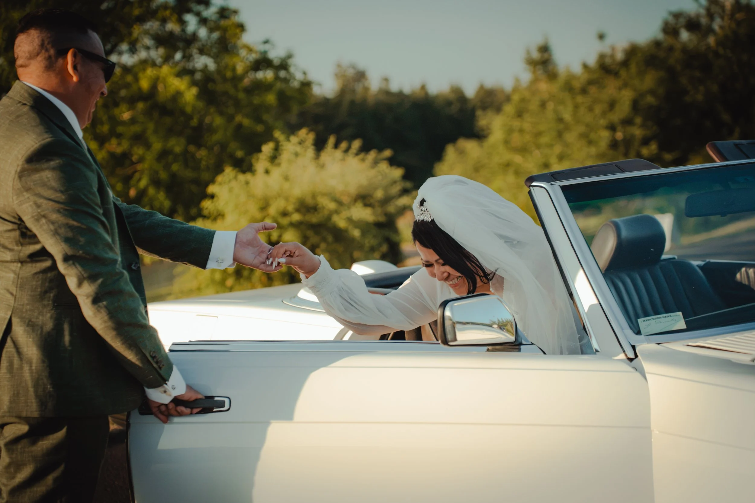 A woman in a wedding dress and veil inside a white convertible car, smiling, as a man in a green suit hands her the keys. The scene is outdoors with trees in the background.