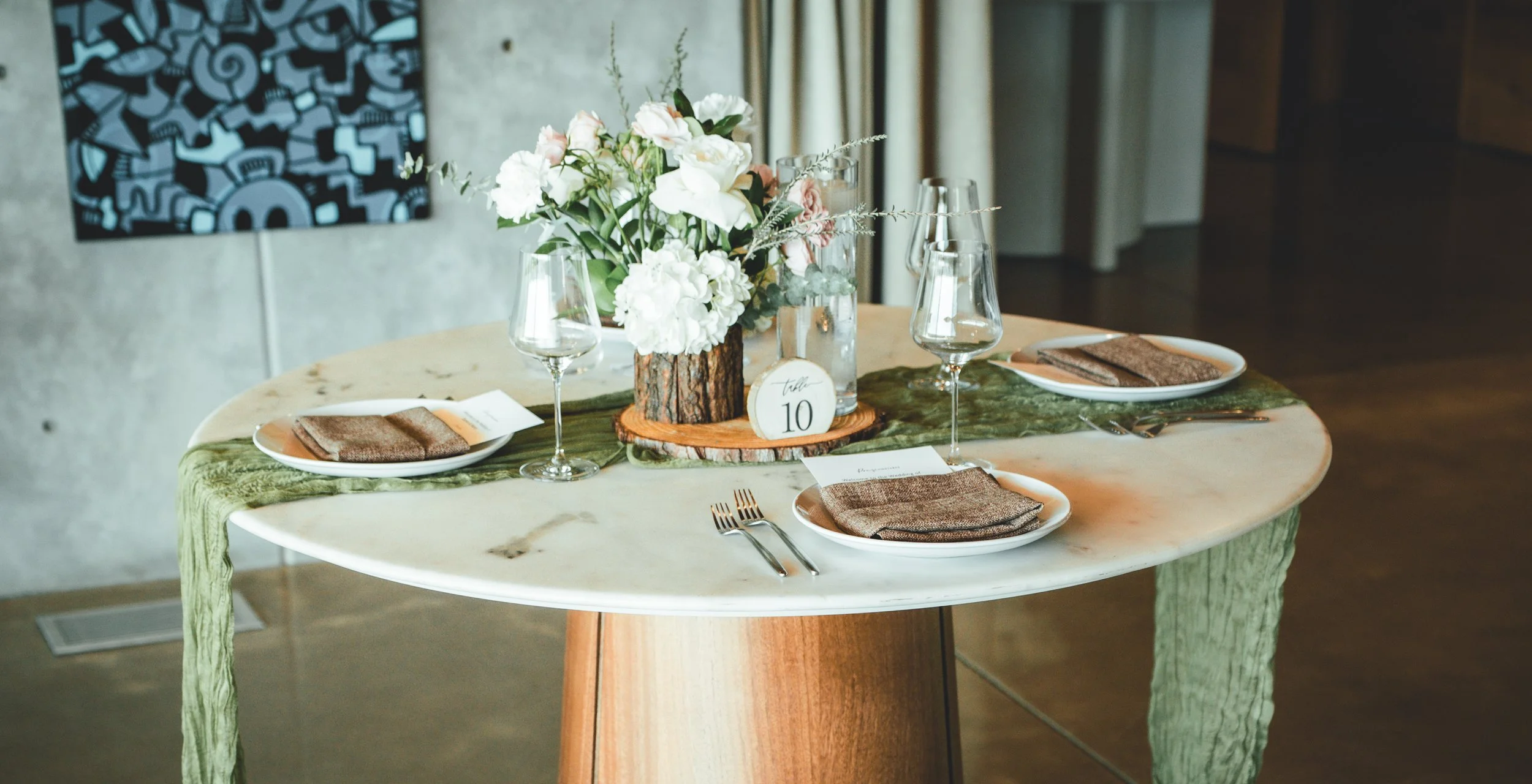 Oval dining table set for a meal with a centerpiece of white and pink flowers in a wood log vase, two wine glasses, plates with napkins, and table number 10, with a green table runner, in a modern room.