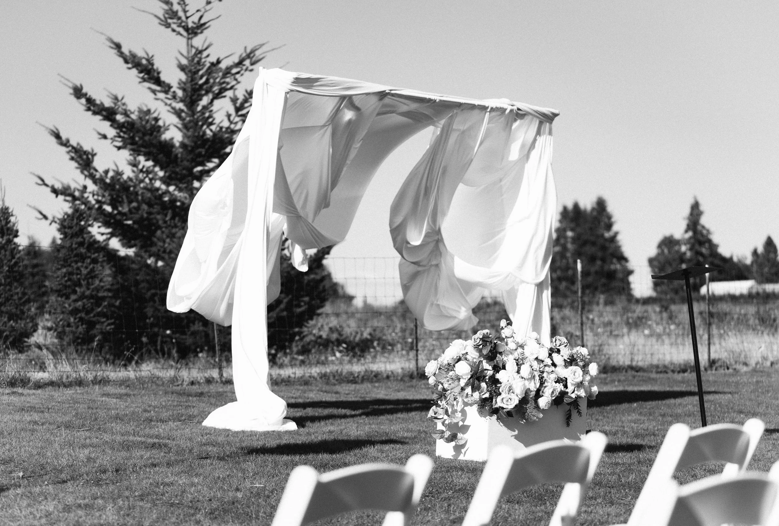 Outdoor wedding setup with floral arrangements, draped fabric, and white chairs on grass, with trees in the background.