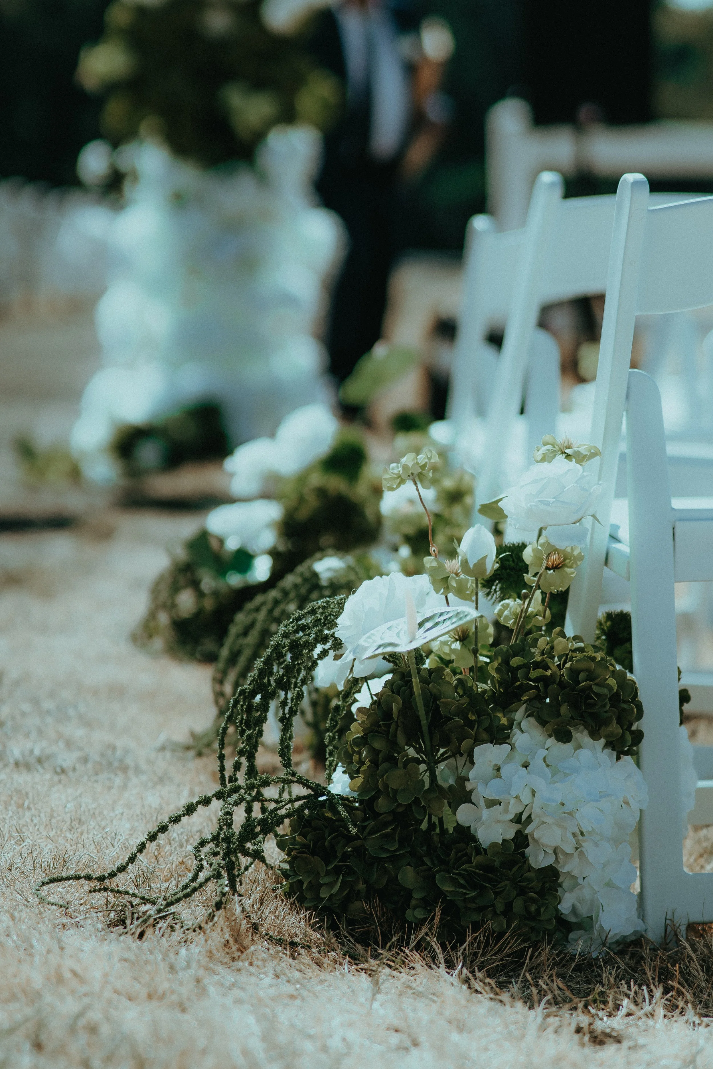 White chairs and floral decorations on a sandy surface, possibly at an outdoor wedding or event setup.