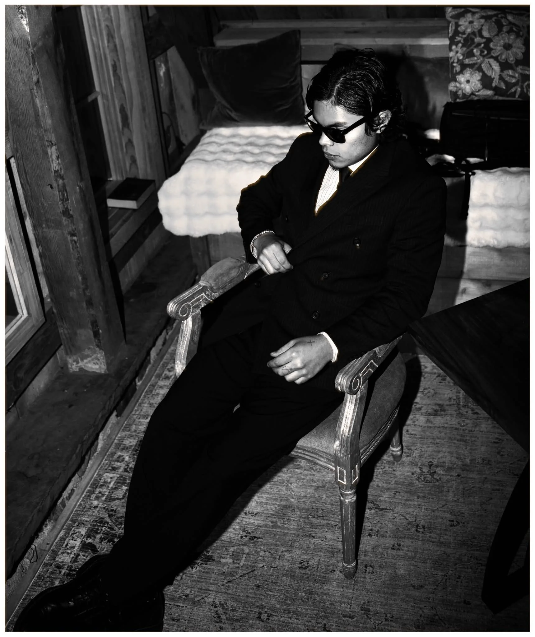 A young man in a black suit and sunglasses sitting on a vintage wooden chair in a room with wooden walls and a patterned carpet.