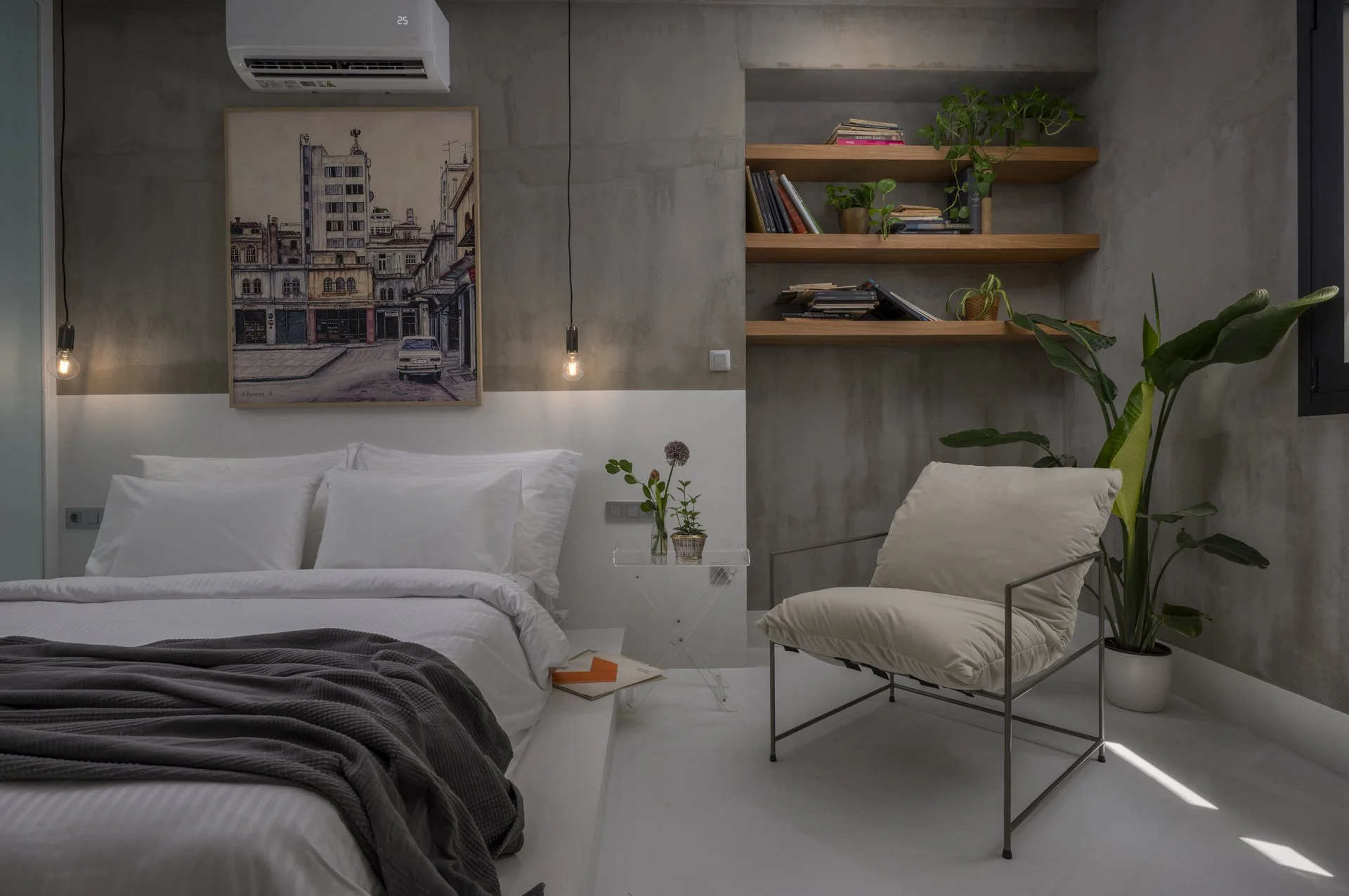 Modern bedroom with a white bed, a gray blanket, a small side table with a plant, a framed cityscape painting, hanging light bulbs, a beige armchair, tall green plants, and wooden shelves with books and decor, set against gray concrete walls.