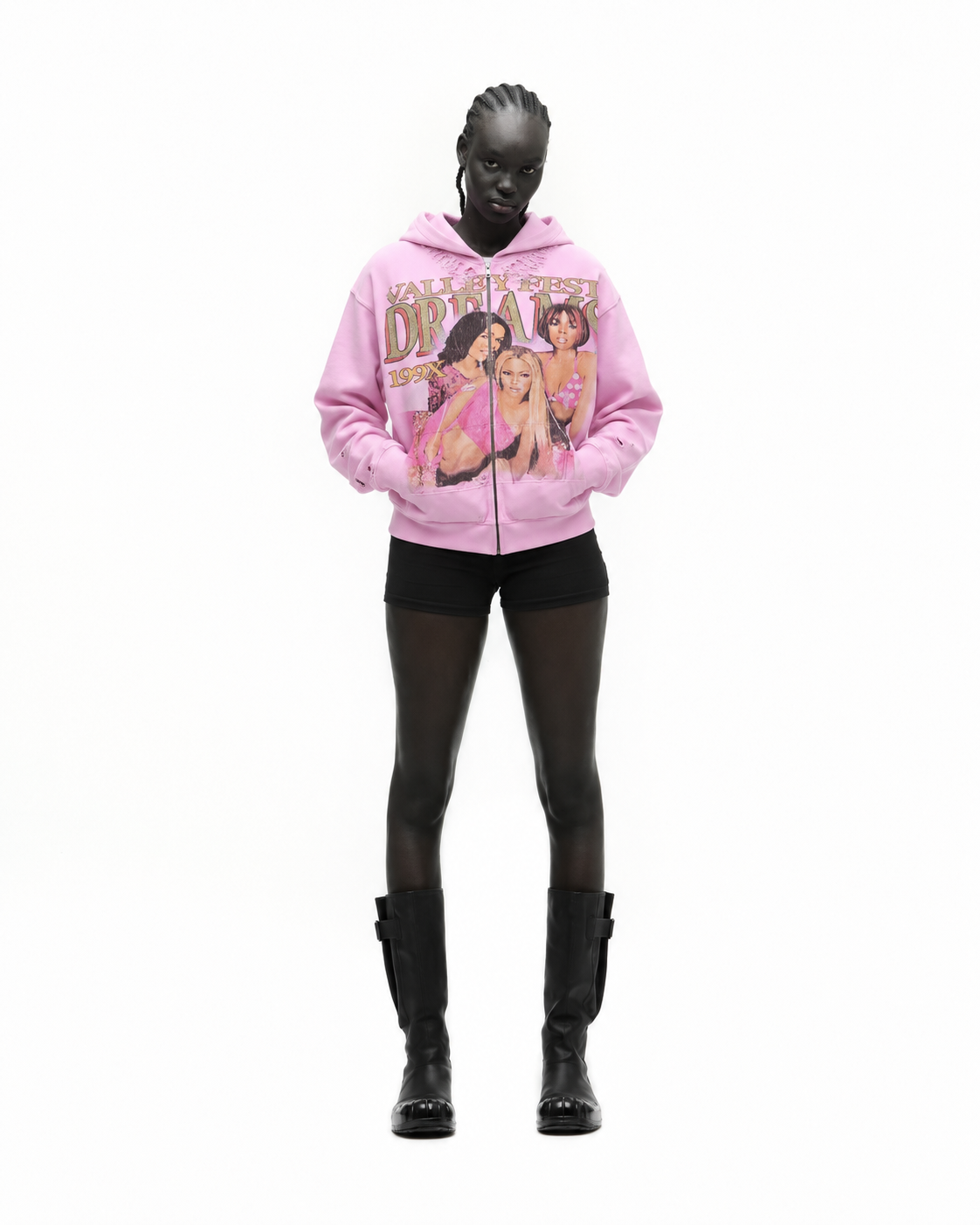 A woman with dark skin and braided hair wearing a pink hoodie with colorful graphic print and black shorts, standing against a white background.