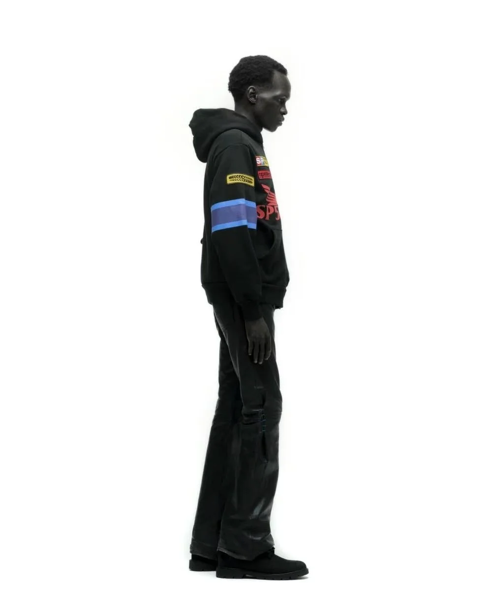 A young man standing in profile against a white background, wearing a black hoodie with colorful patches, black pants, and black shoes.