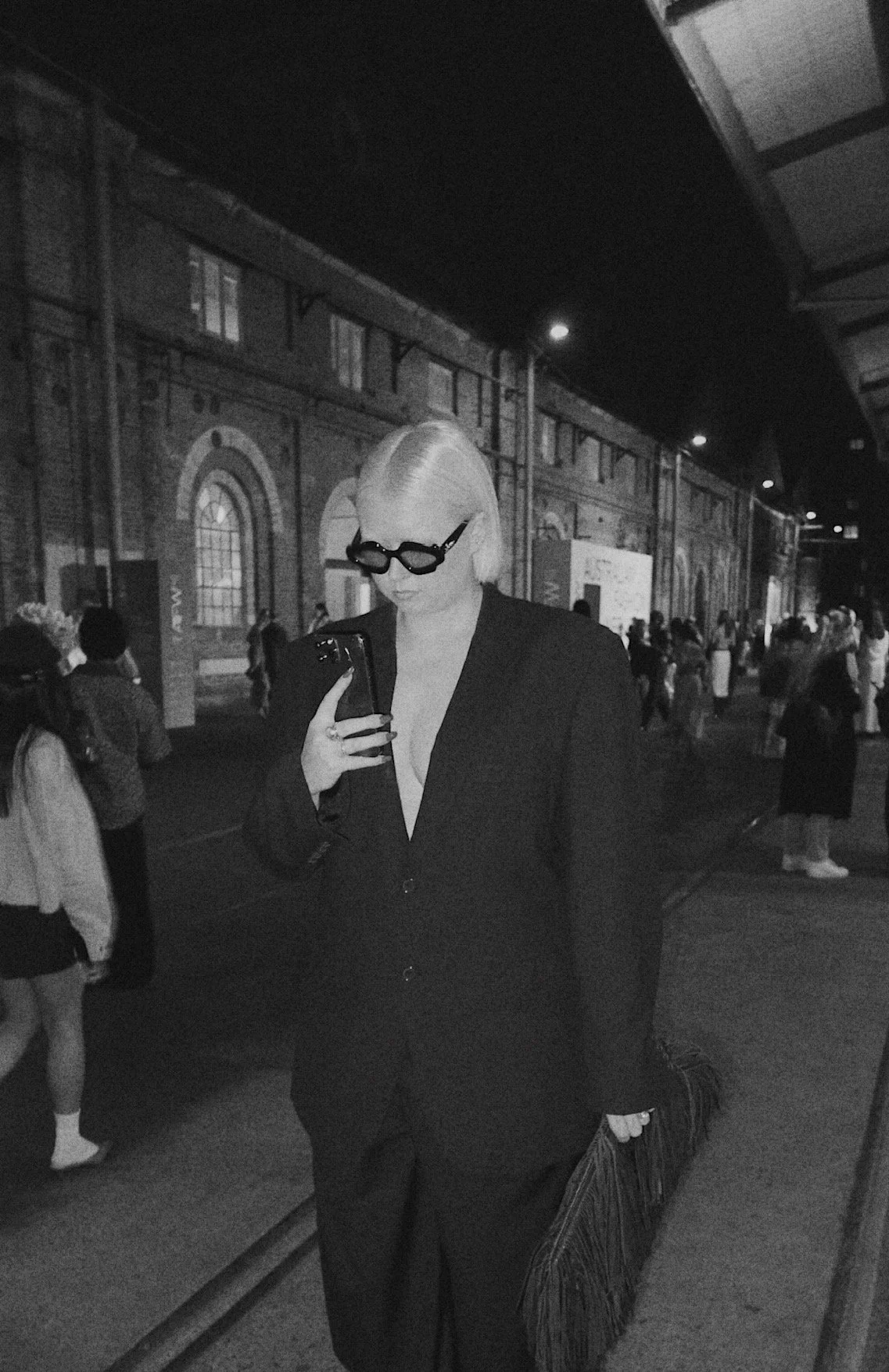 A woman with platinum blonde hair wearing dark sunglasses and a black suit, looking at her phone in a crowded, urban outdoor setting at night.
