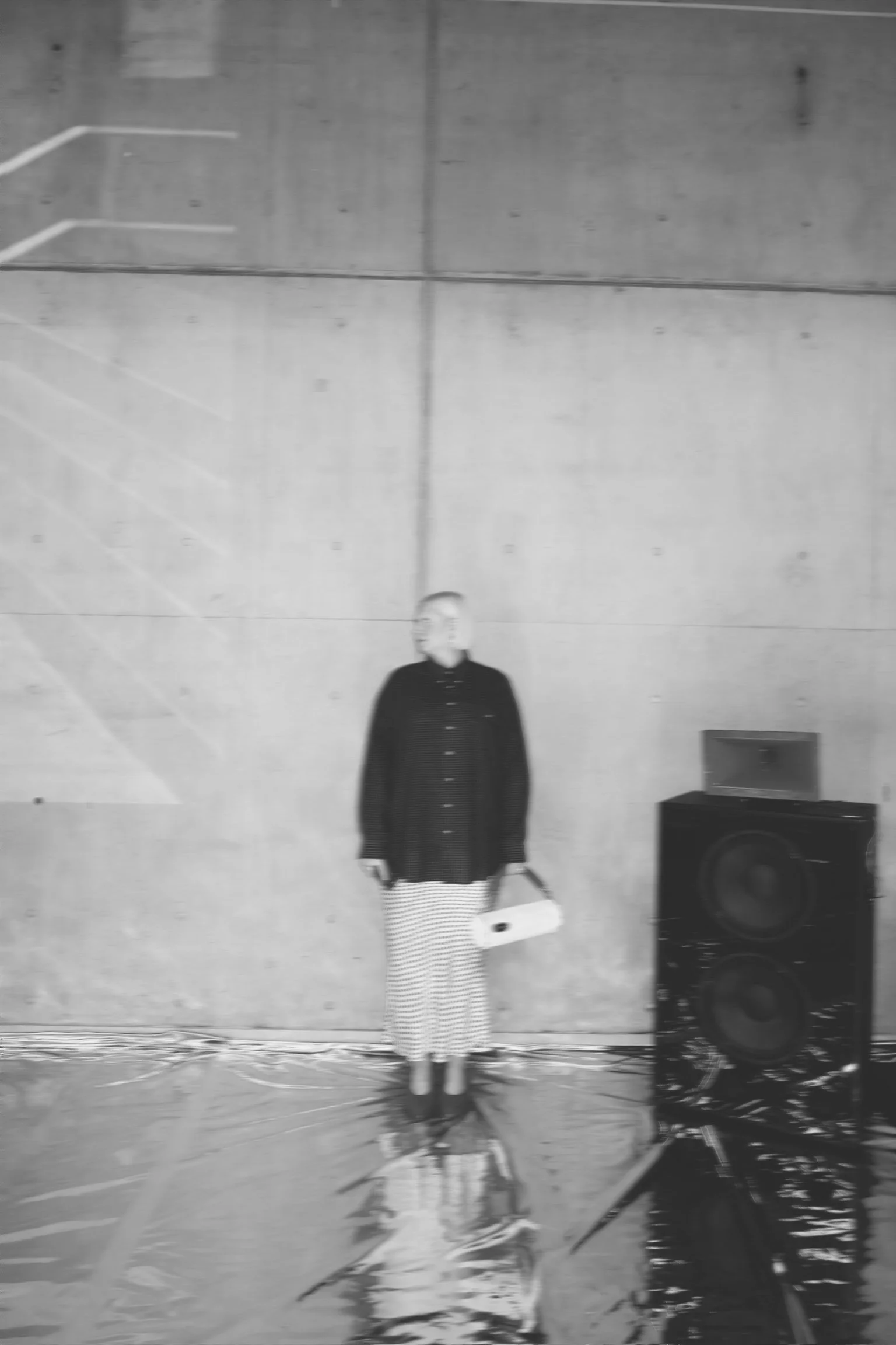 A woman standing in an indoor space with a concrete wall behind her, holding a bag in one hand and a purse in the other, with audio equipment on the floor nearby.