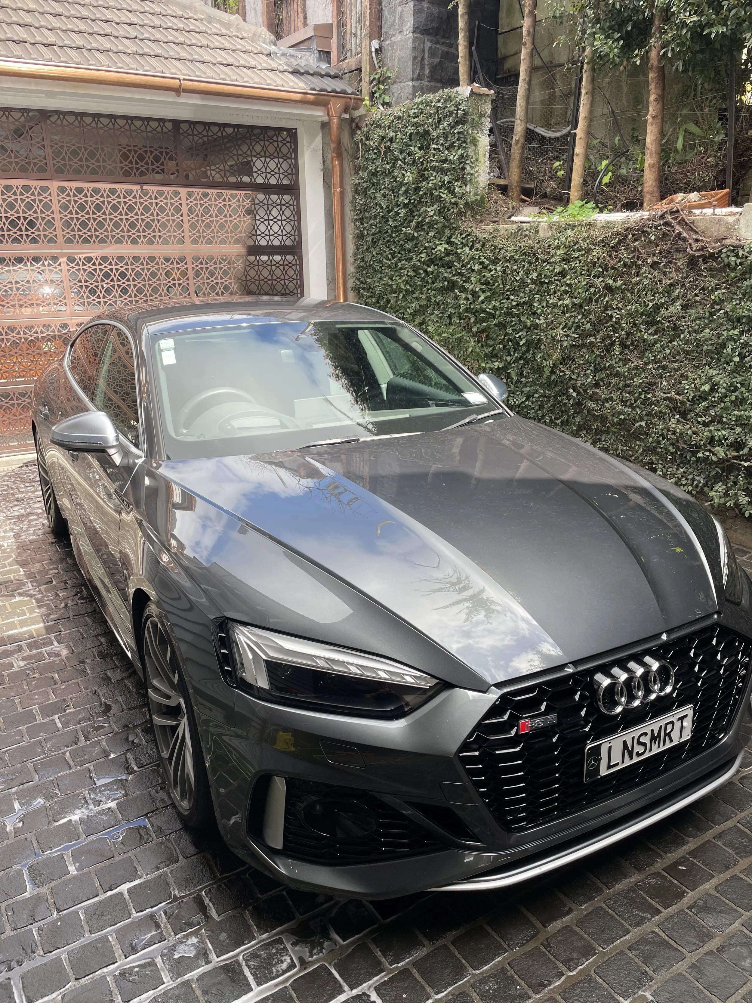A sleek gray Audi RS model car parked on a brick driveway. The front of the car is visible with the Audi logo and license plate reading 'LNSMRT'. Reflective surfaces on the car show the sky and nearby trees. The background includes a house with a gar
