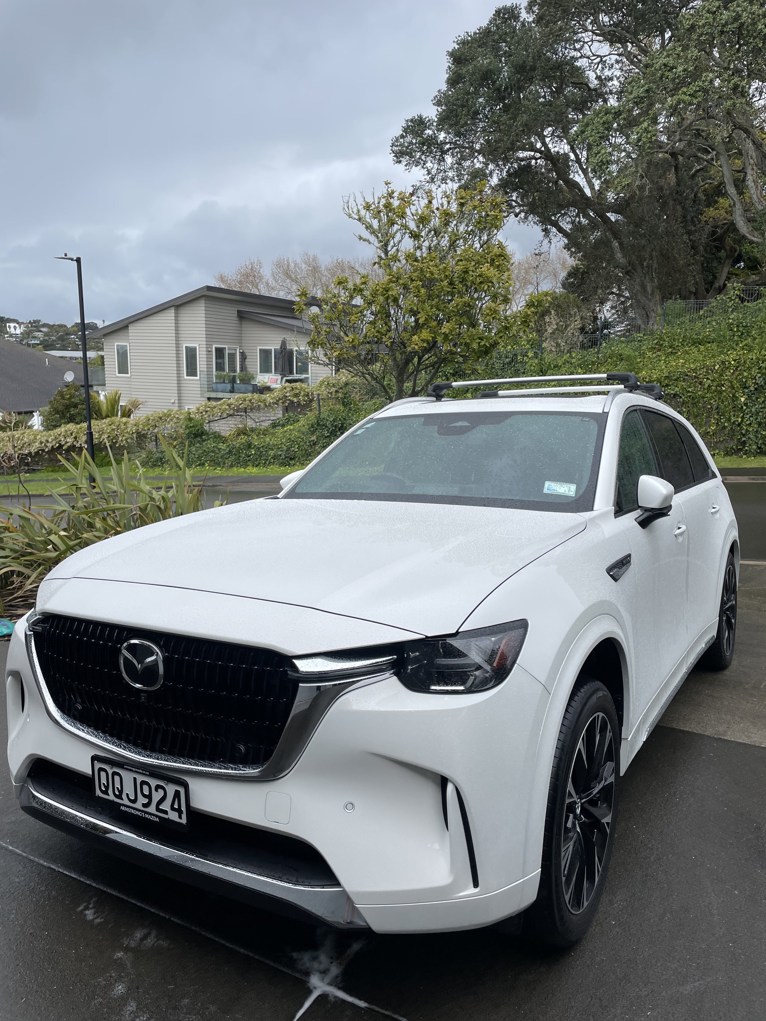 A white Mazda vehicle parked outdoors with a house, trees, and overcast sky in the background.