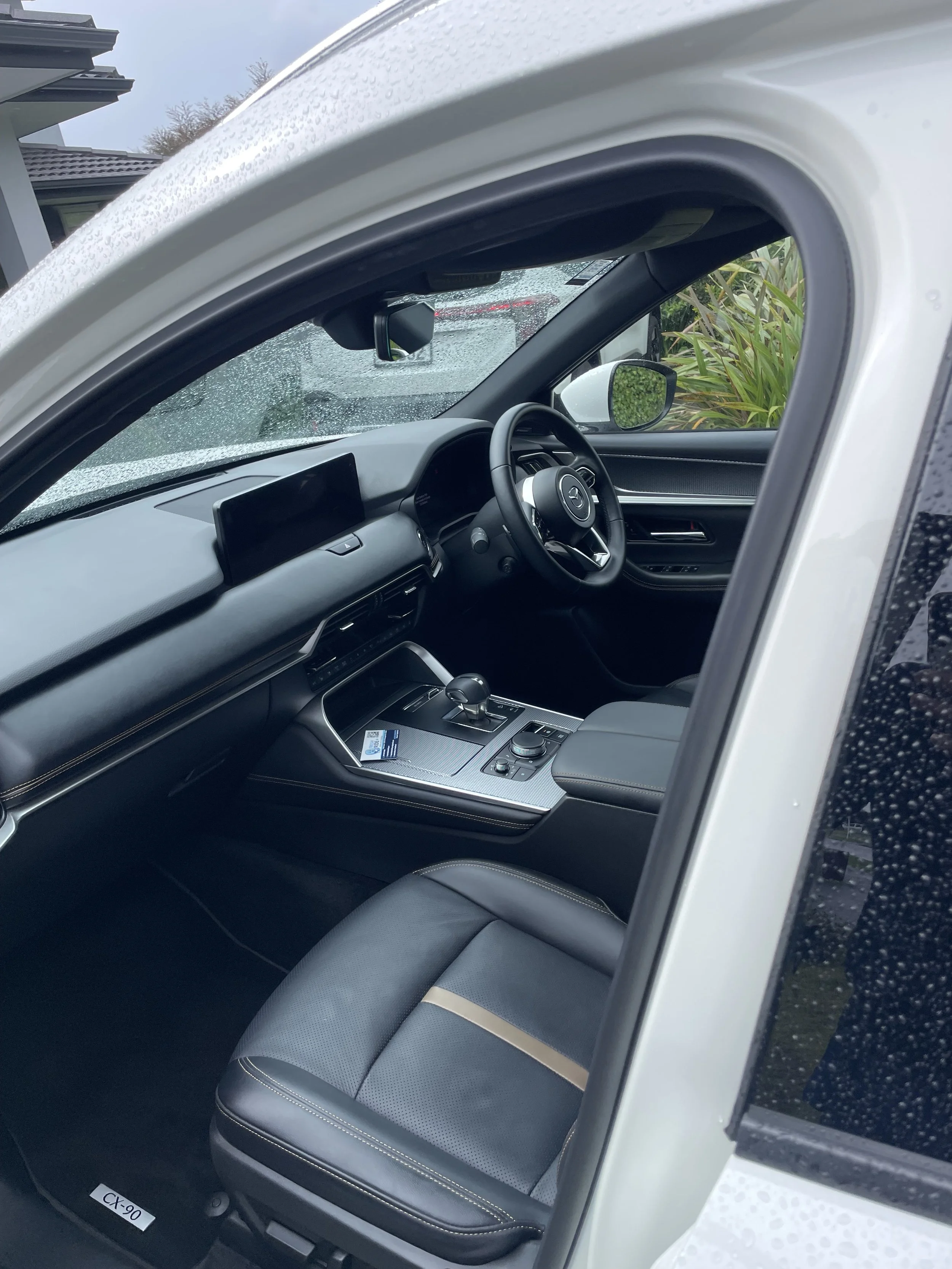 The interior of a modern car showing the driver's seat, dashboard, steering wheel, and center console seen through the open window in rainy weather.