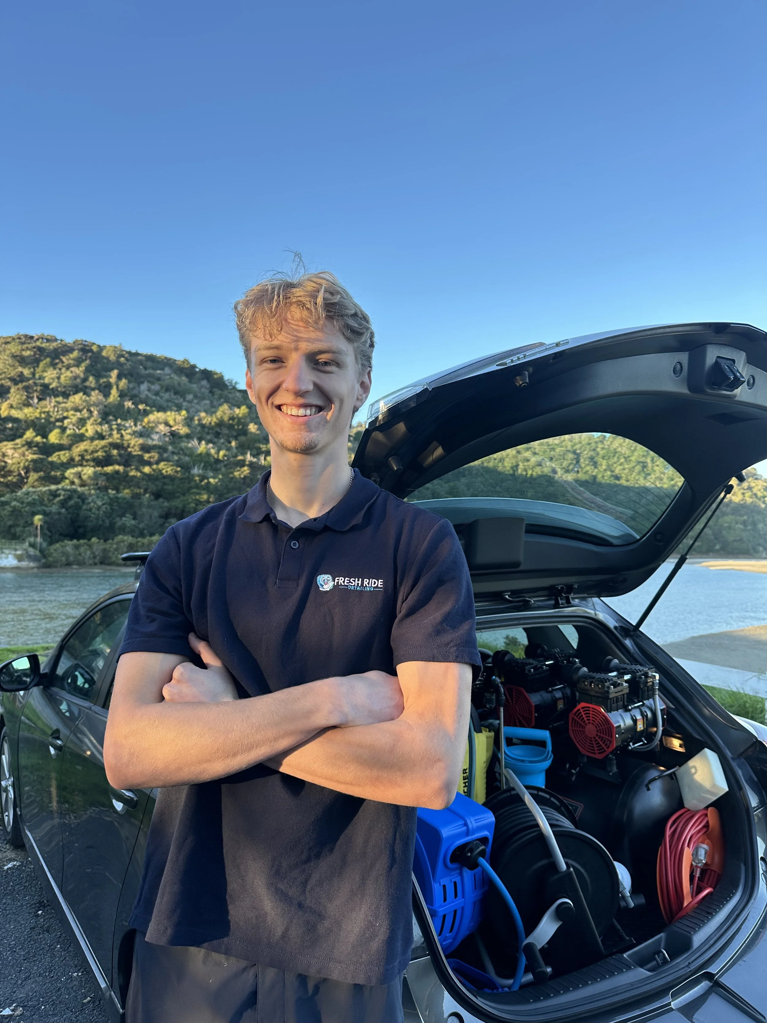 A young man standing with arms crossed in front of an open car trunk, smiling, with tools and equipment inside the trunk, greenery and hills in the background, clear blue sky overhead.