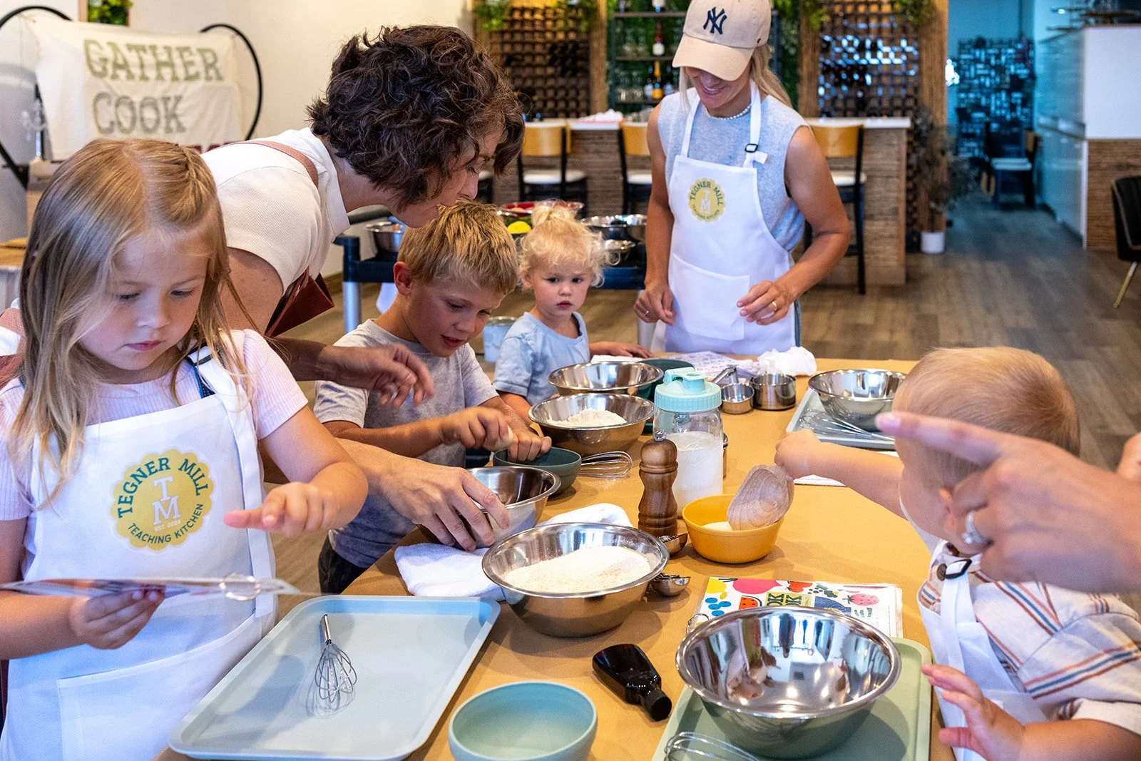 Kids and adults cooking in a kitchen classroom, wearing aprons, surrounded by bowls, utensils, and ingredients.