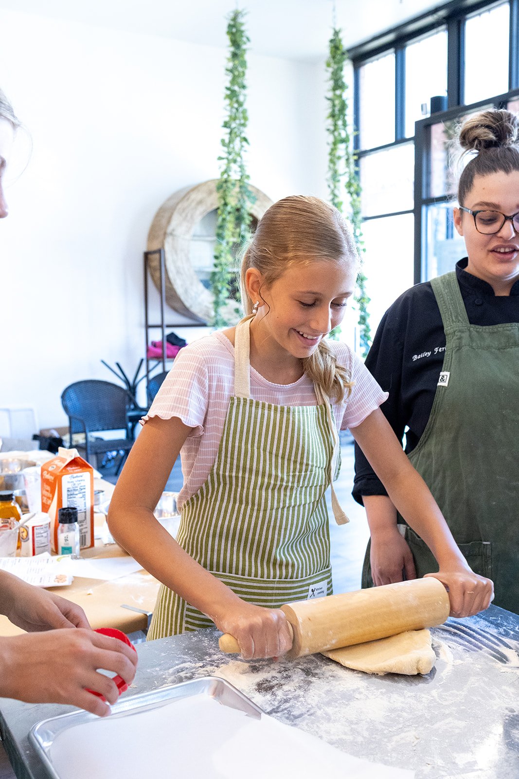 A young girl smiling while rolling dough with a rolling pin in a kitchen or bakery, with a woman standing next to her, in a cooking or baking class.