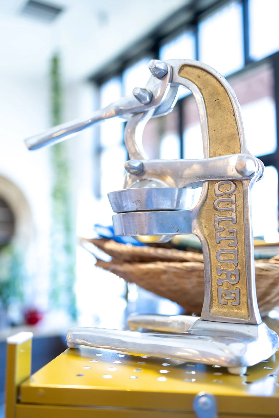 A silver and gold lemon juicer on a yellow surface with a woven basket in the background.