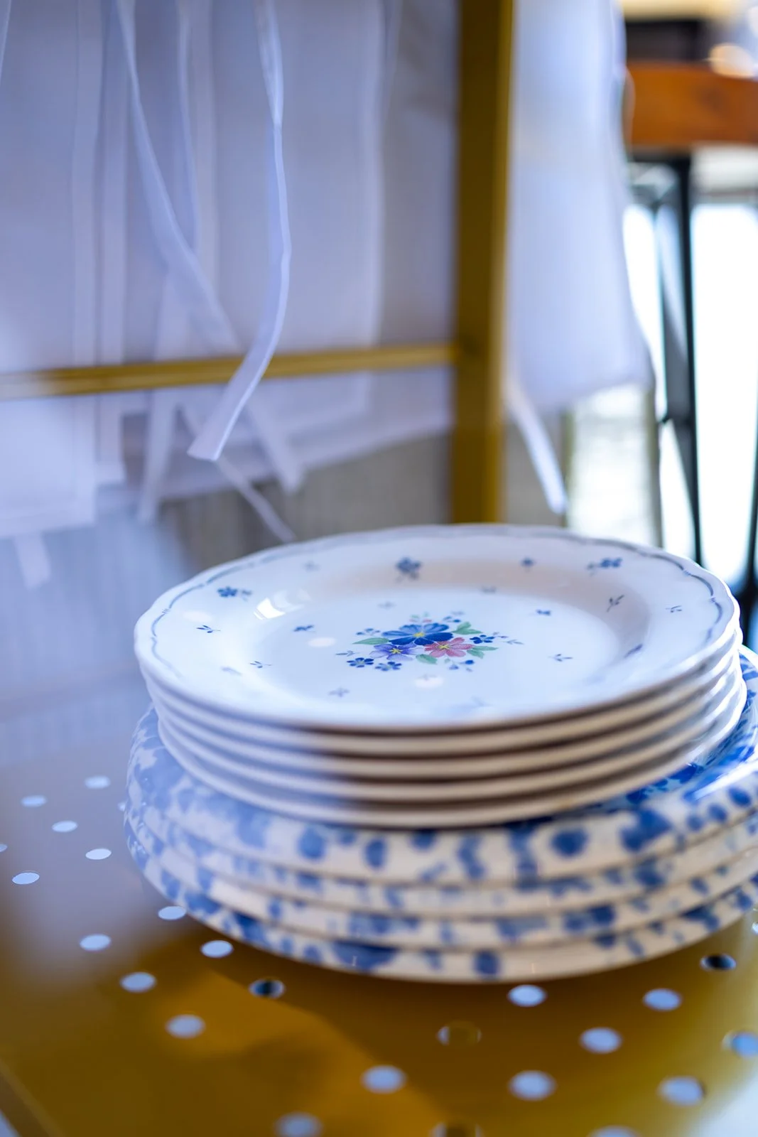 Stack of floral patterned plates on a perforated yellow metal surface, with a background of a wooden chair and hanging white fabric or curtain.
