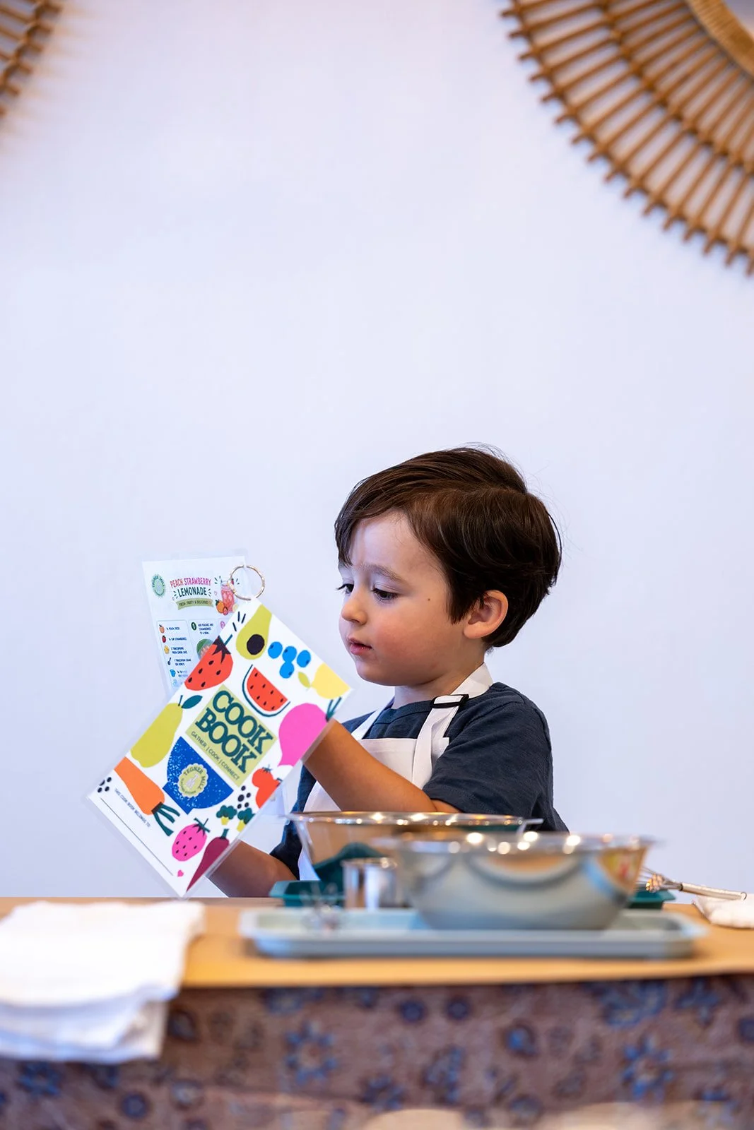 A young boy with brown hair wearing a dark shirt and white apron looking at a colorful cookbook at a kitchen counter.