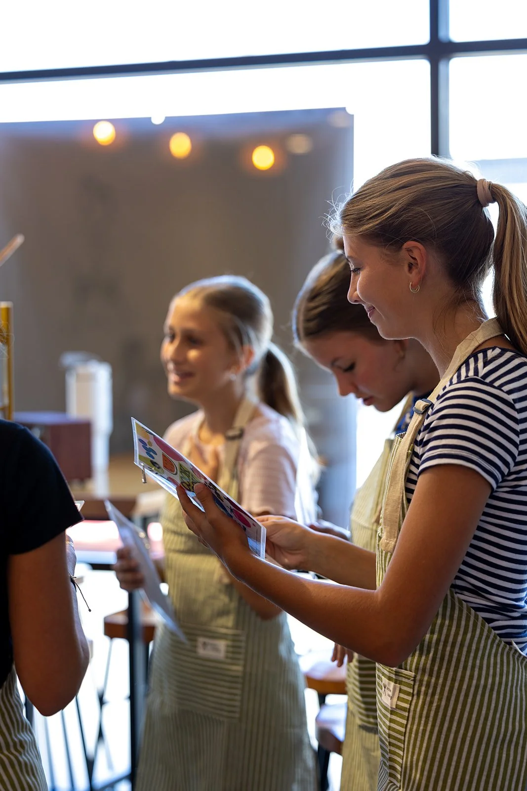 Group of girls wearing aprons in a kitchen, looking at a recipe or instructions, smiling and engaged in cooking or baking activity.