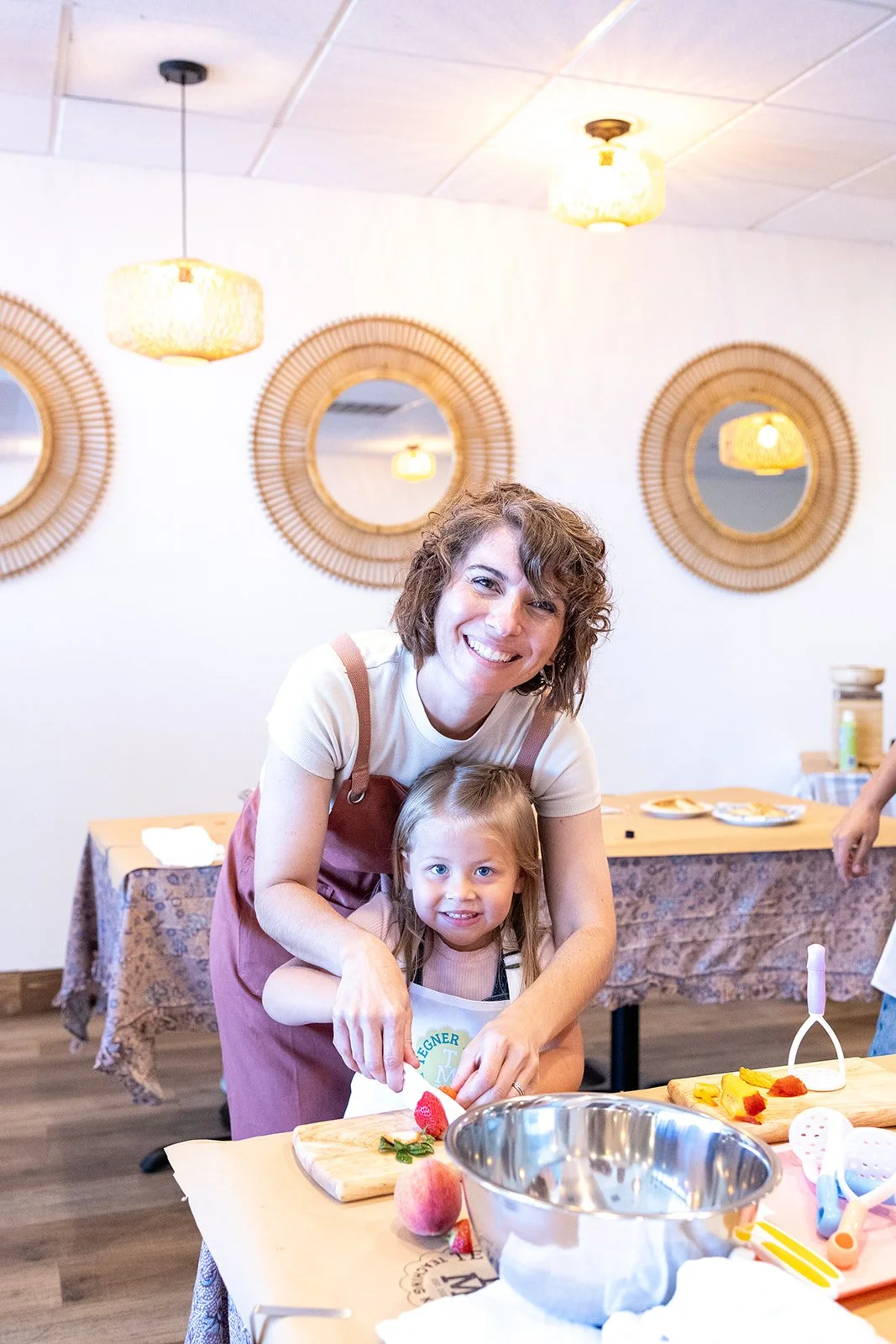 A woman and a young girl smiling while preparing food together in a kitchen with decorative round mirrors on the wall and warm lighting.