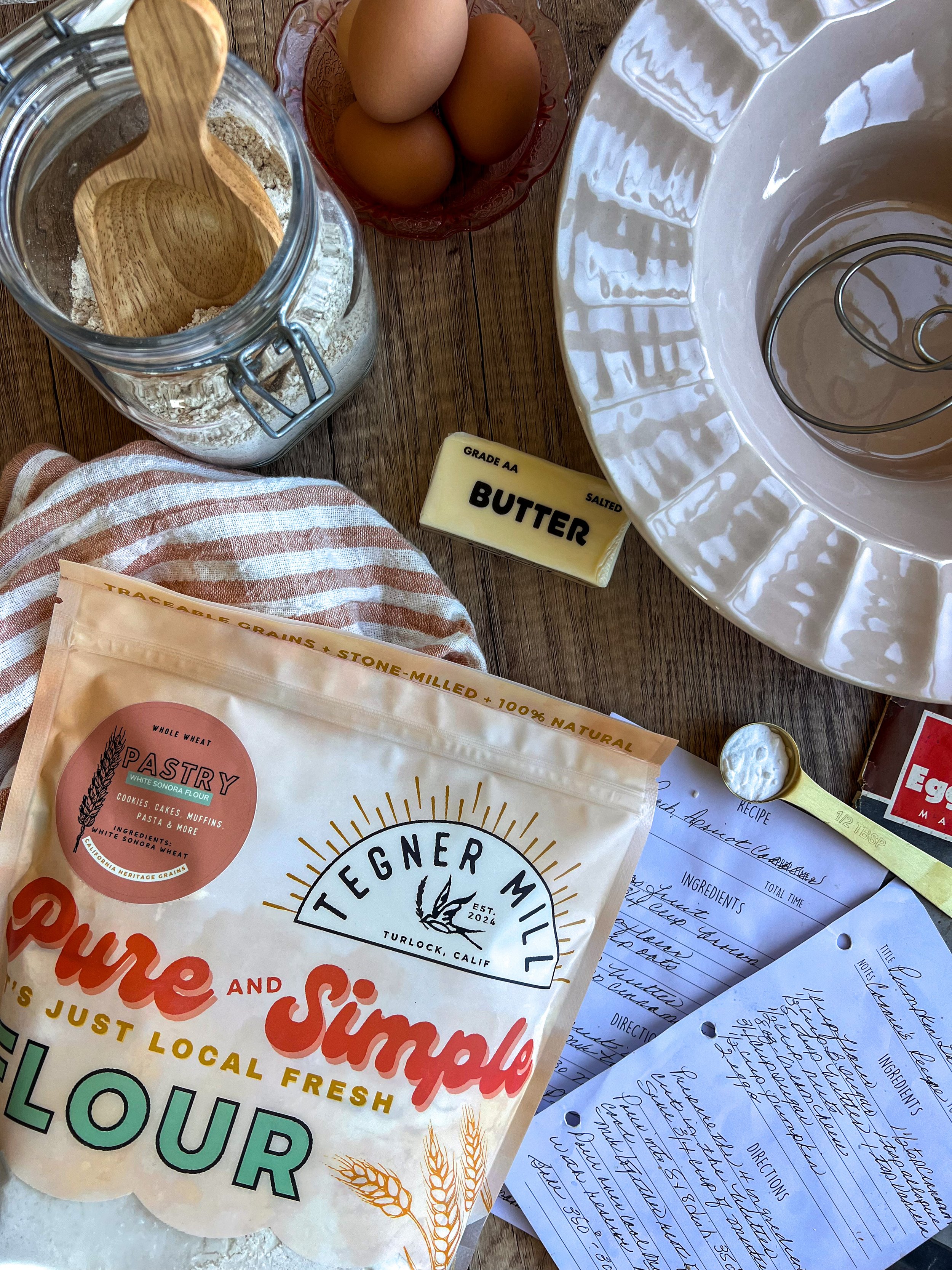 A collection of baking ingredients and tools on a wooden surface, including a bag of flour, a stick of salted butter, a jar with a wooden spoon, a bowl of eggs, a wire whisk inside a fluted ceramic mixing bowl, and handwritten recipe notes.