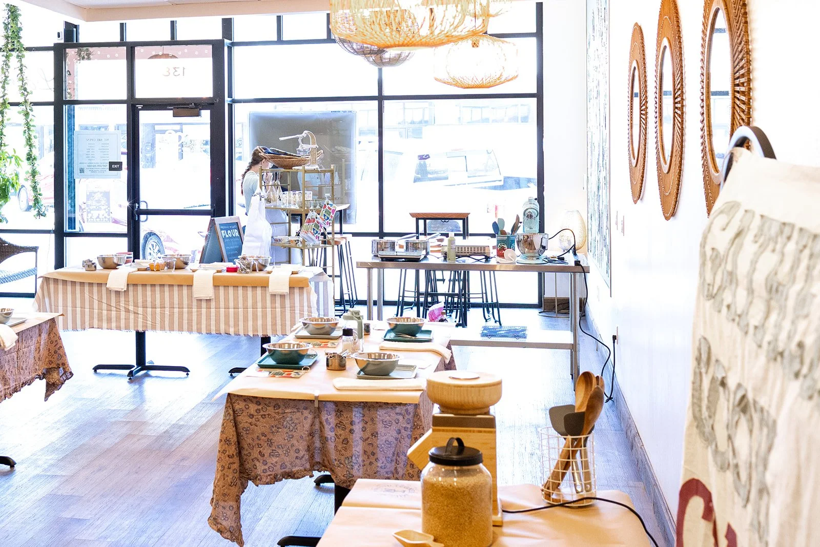 Interior of teaching kitchen with tables set with bowls, plates, and utensils, decorated with patterned tablecloths, with large windows allowing natural light, and decorative wall mirrors.