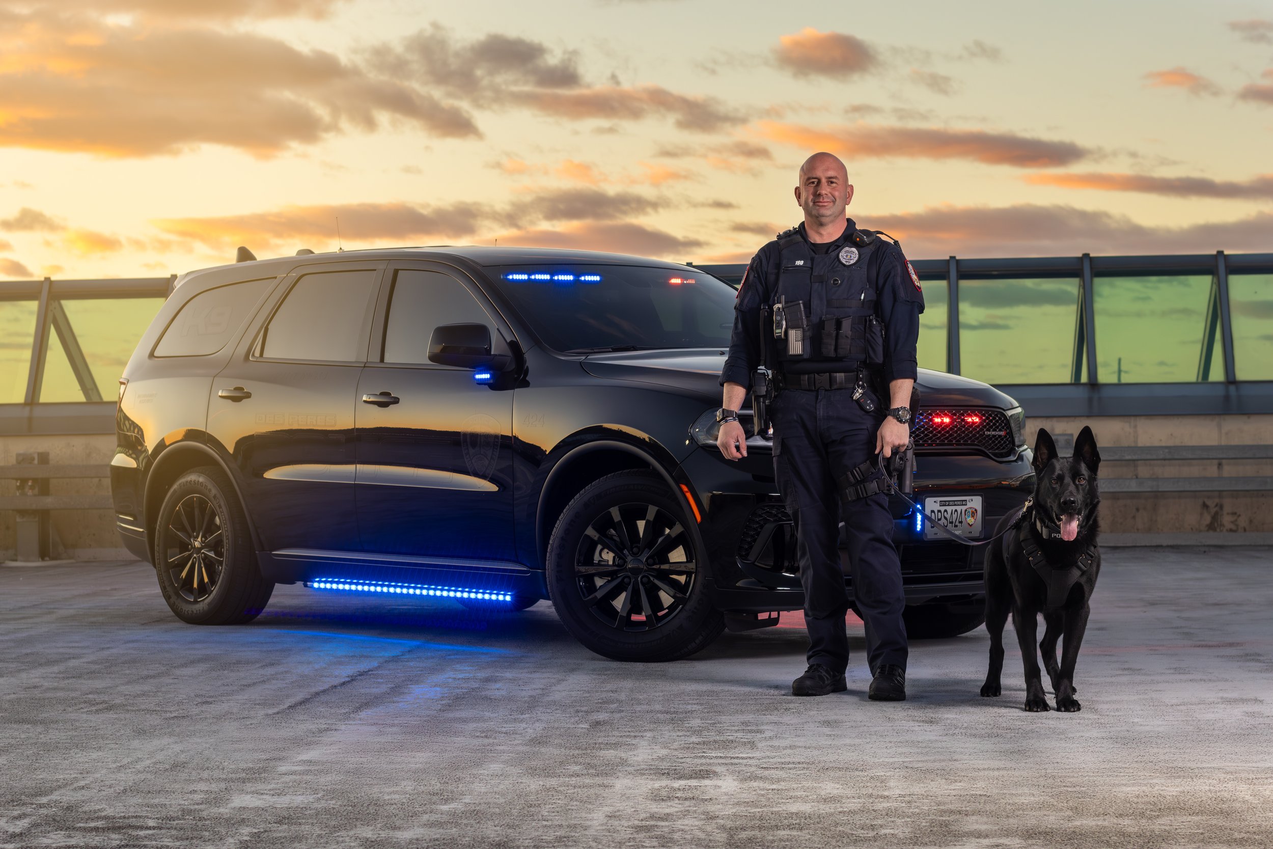 A police officer standing next to a police dog and vehicle on a parking deck during sunset.