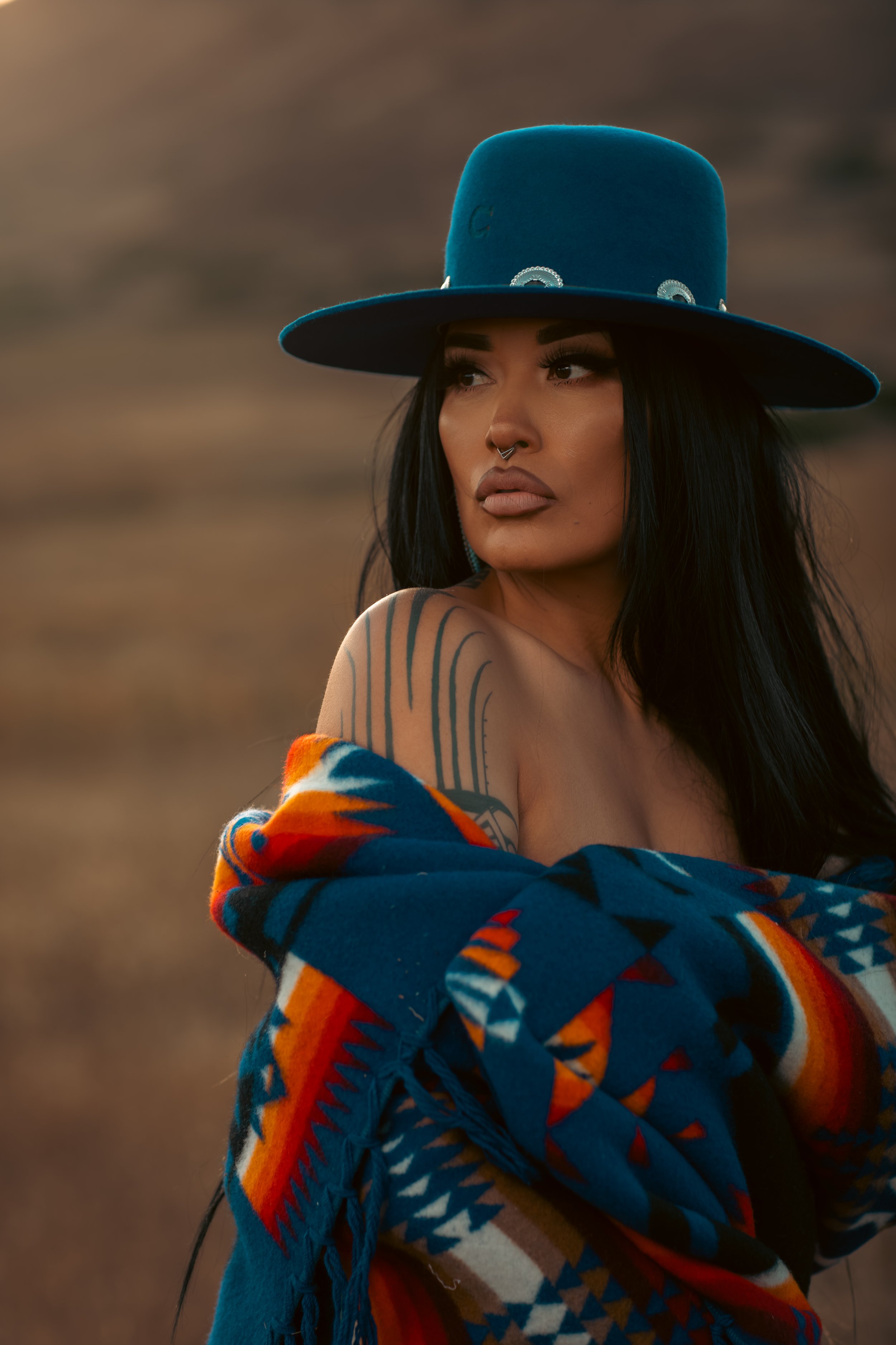 A woman with dark hair, wearing makeup, a septum piercing, and a colorful patterned blanket or shawl, is outdoors with a blurred natural background, wearing a large blue hat.