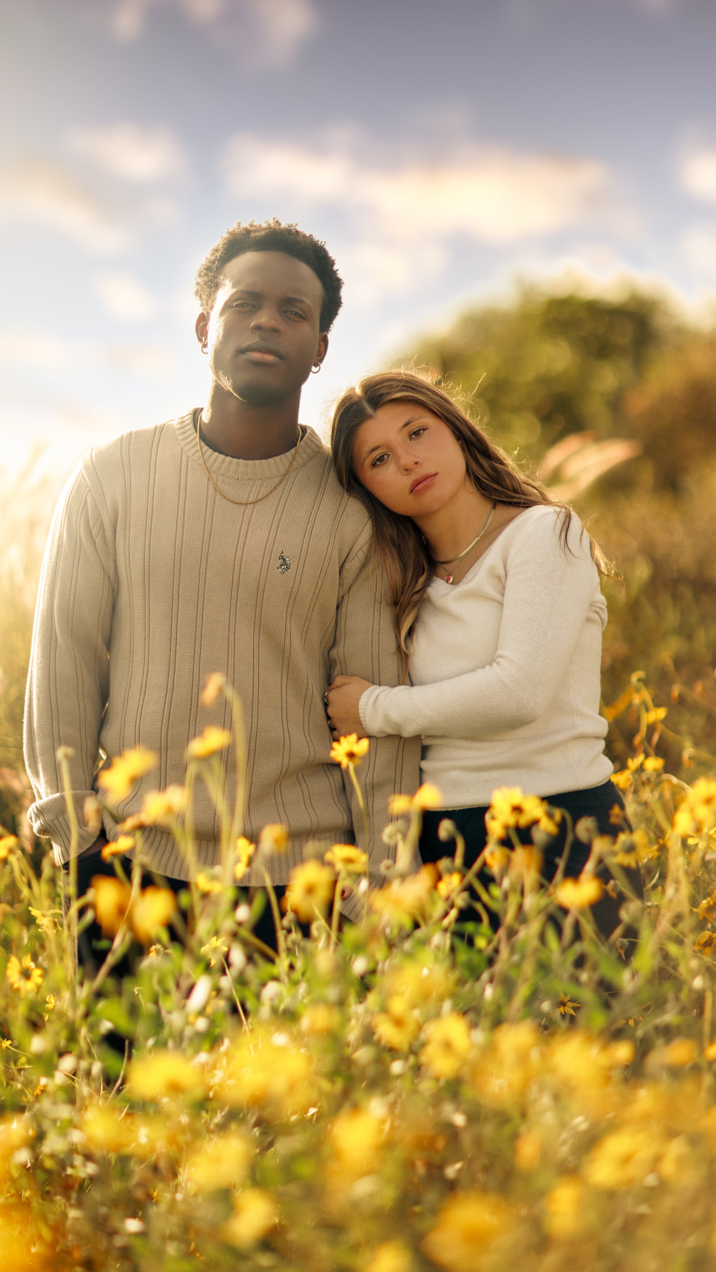 A young man and woman standing close together in a field of yellow flowers during golden hour sunset, with a blurred background of trees and sky.