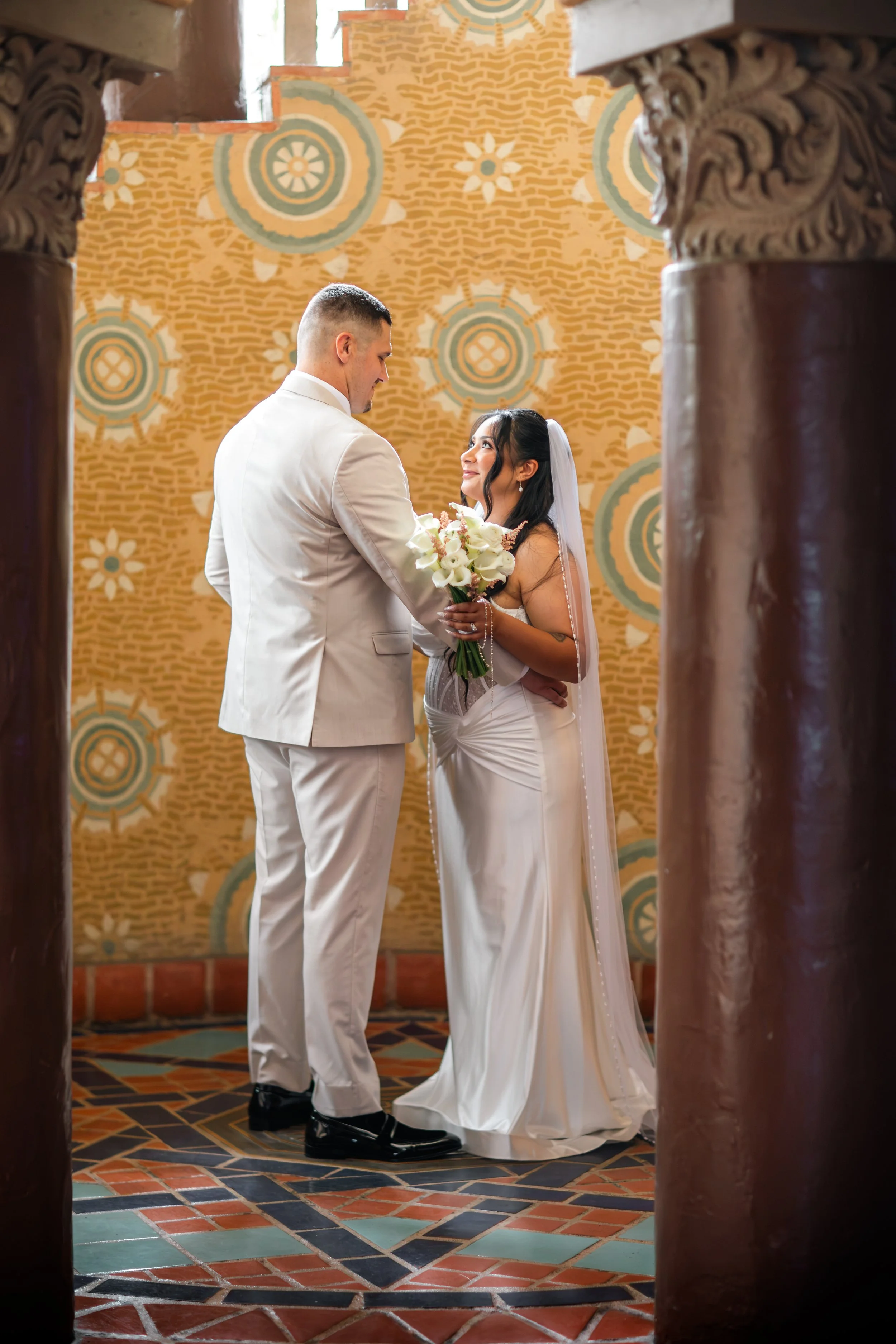 A bride and groom share a moment during their wedding ceremony, framed by ornate wooden columns, with a patterned yellow and green background.