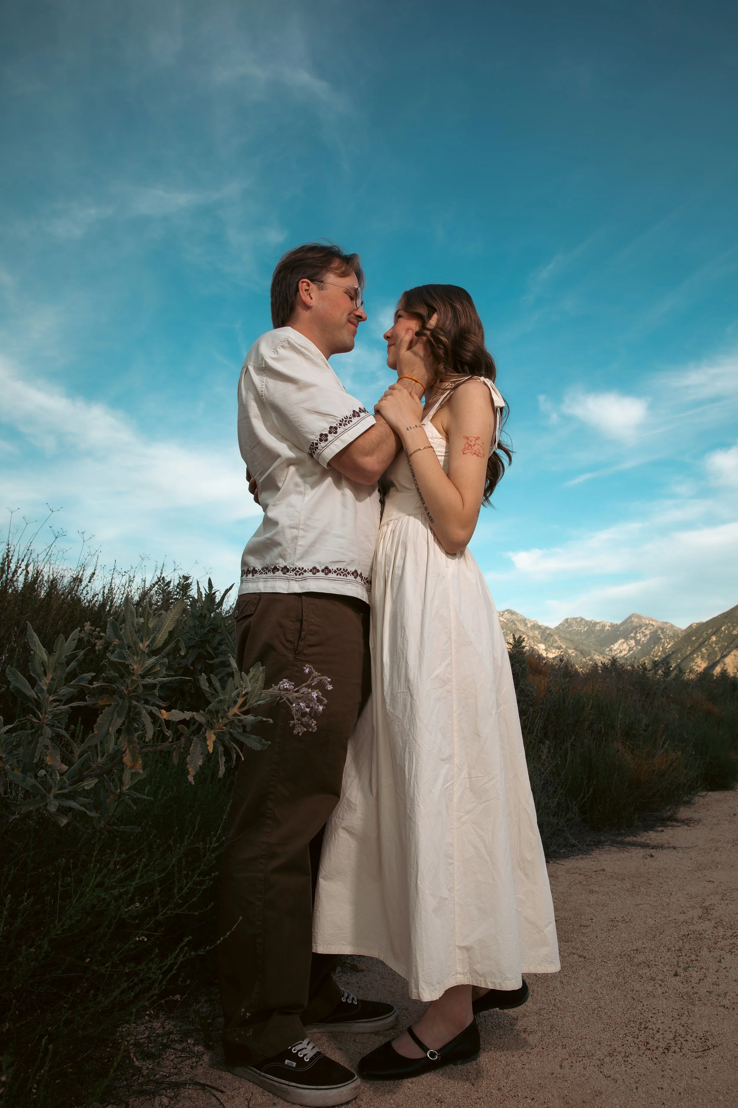 A couple standing close together outdoors against a backdrop of mountains and blue sky, looking at each other lovingly, with the woman gently holding the man's face.