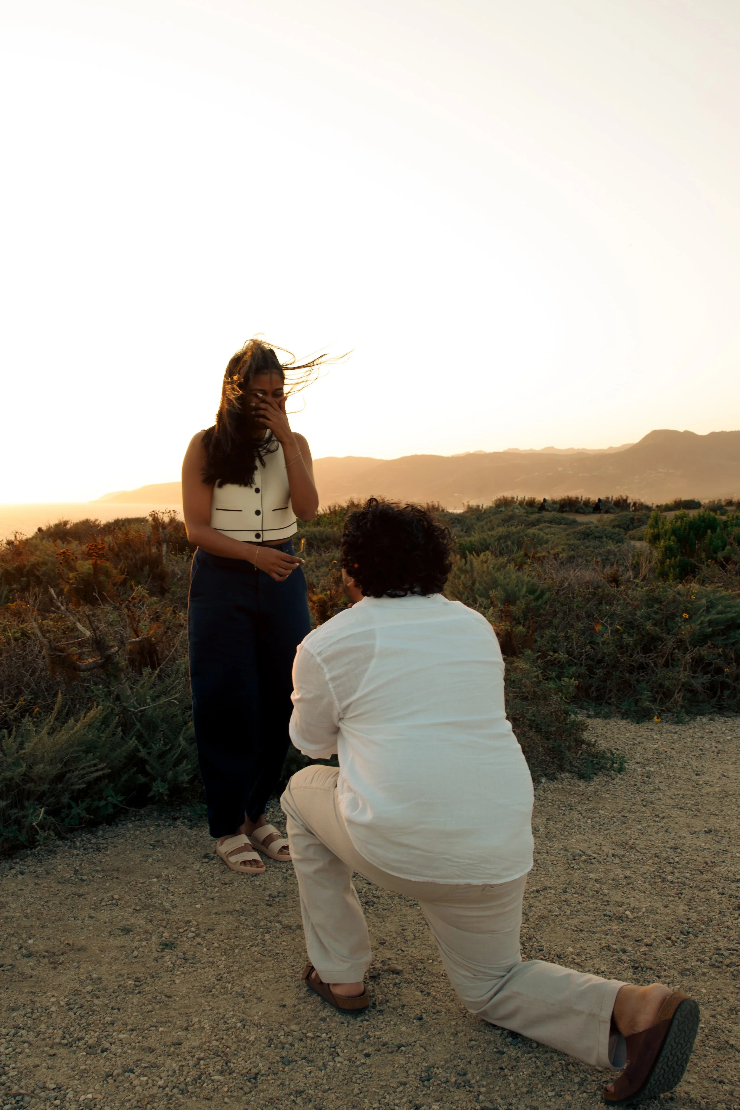 A woman standing with her hand over her mouth, laughing or smiling, while a man kneels in front of her on a gravel trail during sunset, with a mountain range and shrubs in the background.