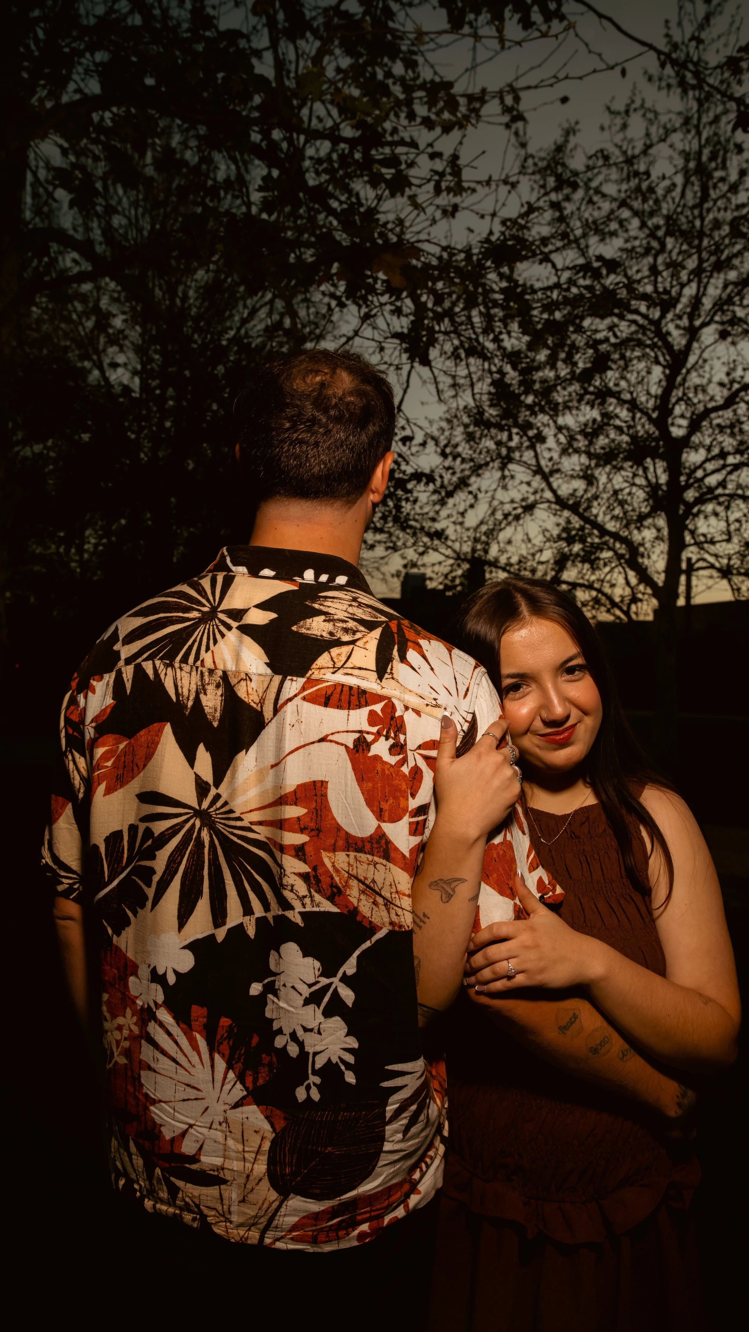 A man with short dark hair wearing a large flower pattern shirt stands close to a woman with long dark hair, wearing a sleeveless brown dress. The man is facing away from the camera, and the woman is smiling at the camera, with her head leaning on hi