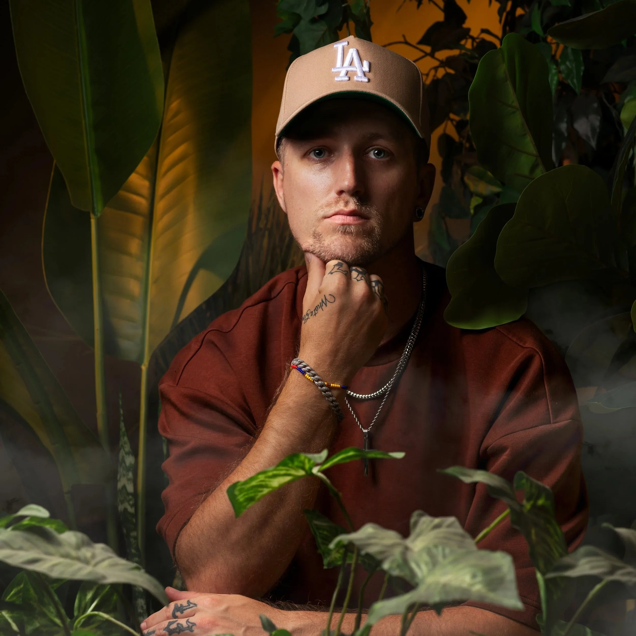 A man with tattoos on his fingers wearing a tan baseball cap with a white 'LA' logo, surrounded by green plants and leaves.