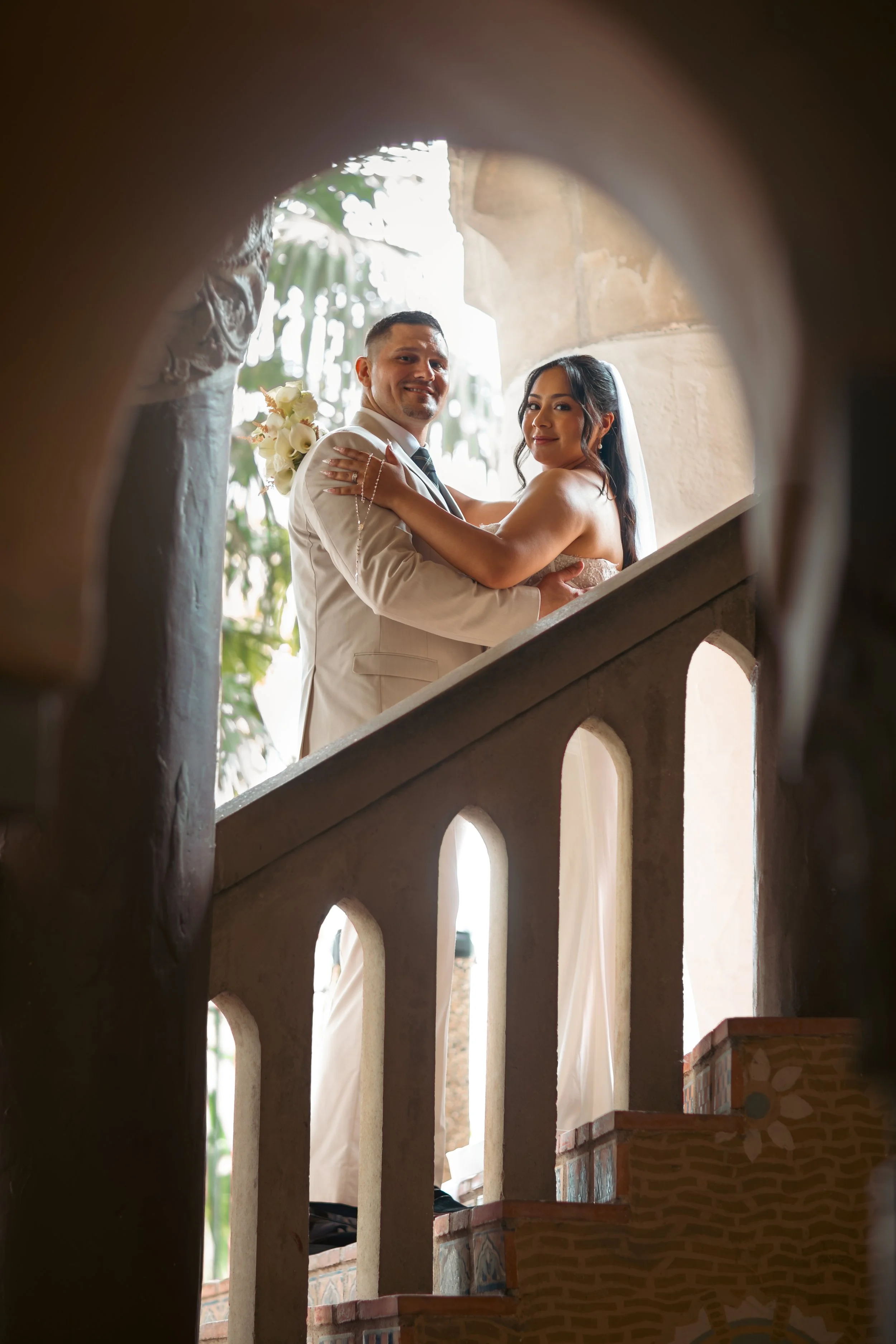 A newlywed couple on a staircase looking through a circular opening, with the groom in a white suit and the bride in a strapless wedding dress, holding a bouquet of white flowers.