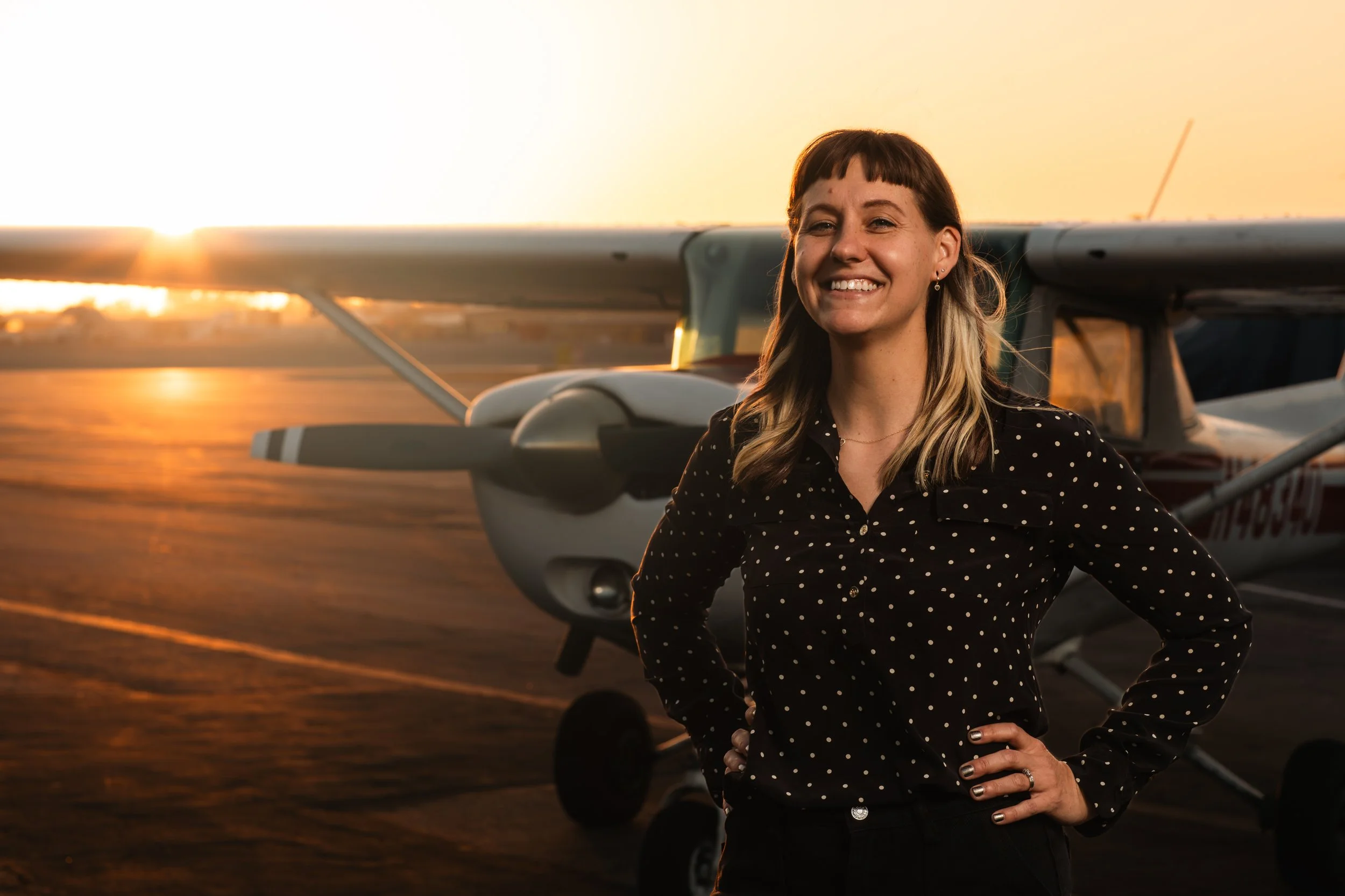 A woman with brown hair and bangs, wearing a black polka dot shirt, smiling and standing in front of a small airplane at sunset.