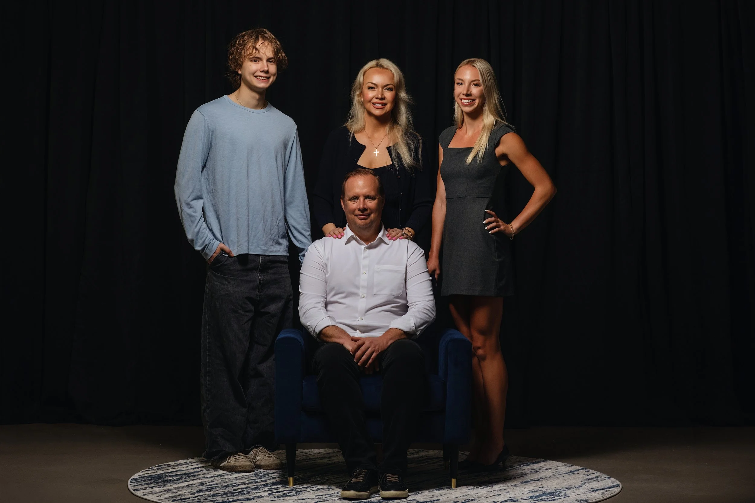 A group of five people posing together on a stage or in a studio with black curtains and a circular rug. One man is seated in a blue chair, surrounded by four women standing behind him.