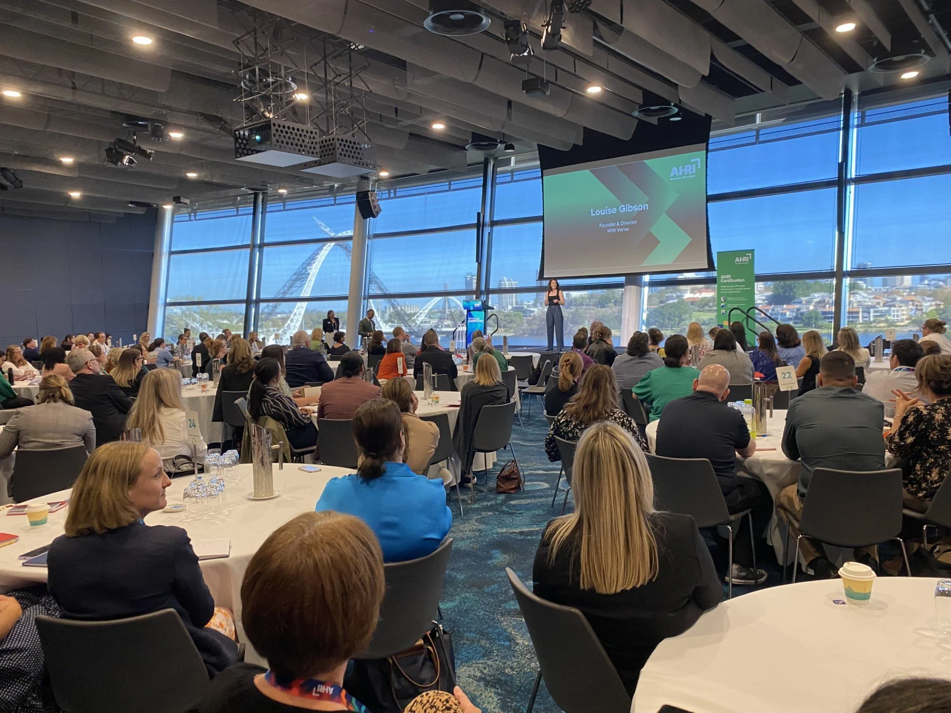 A conference room filled with attendees seated at round tables, listening to a standing woman presenting on stage. Large windows in the background reveal a cityscape and the Eiffel Tower. A large screen displays the speaker's name, Louise Gibson, and the event's theme.