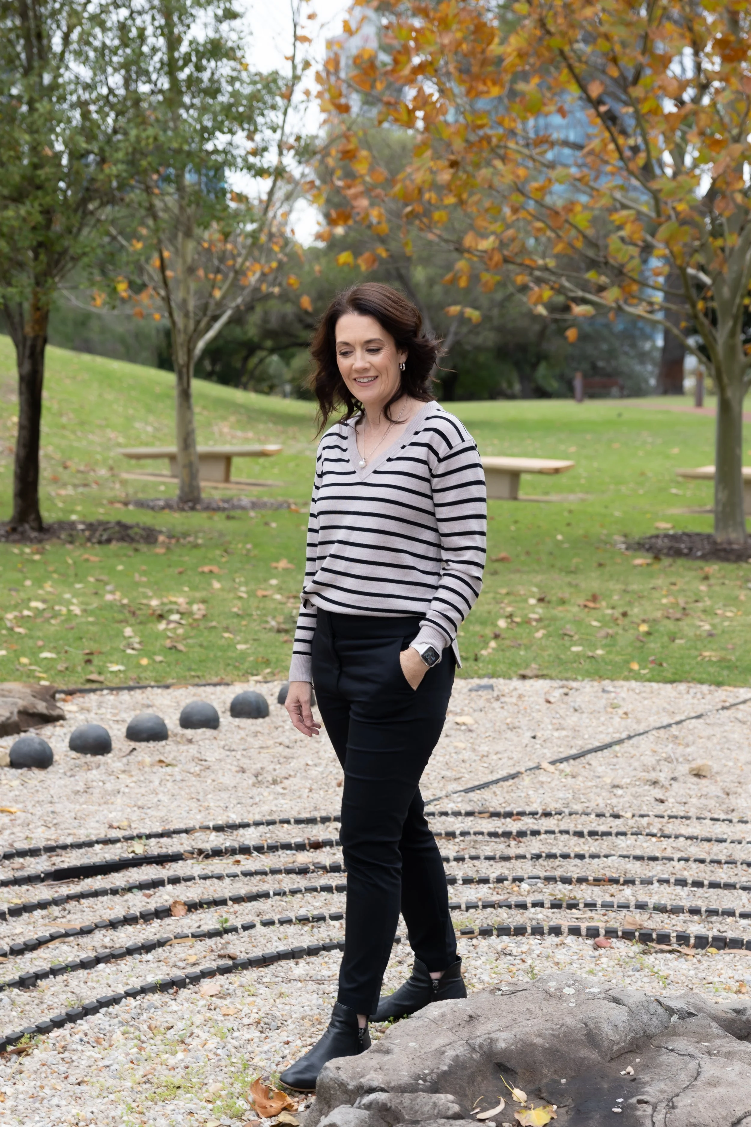 Woman with dark brown hair wearing a striped sweater and black pants standing outdoors in a park with autumn leaves, trees, benches, and rock formation in the background.
