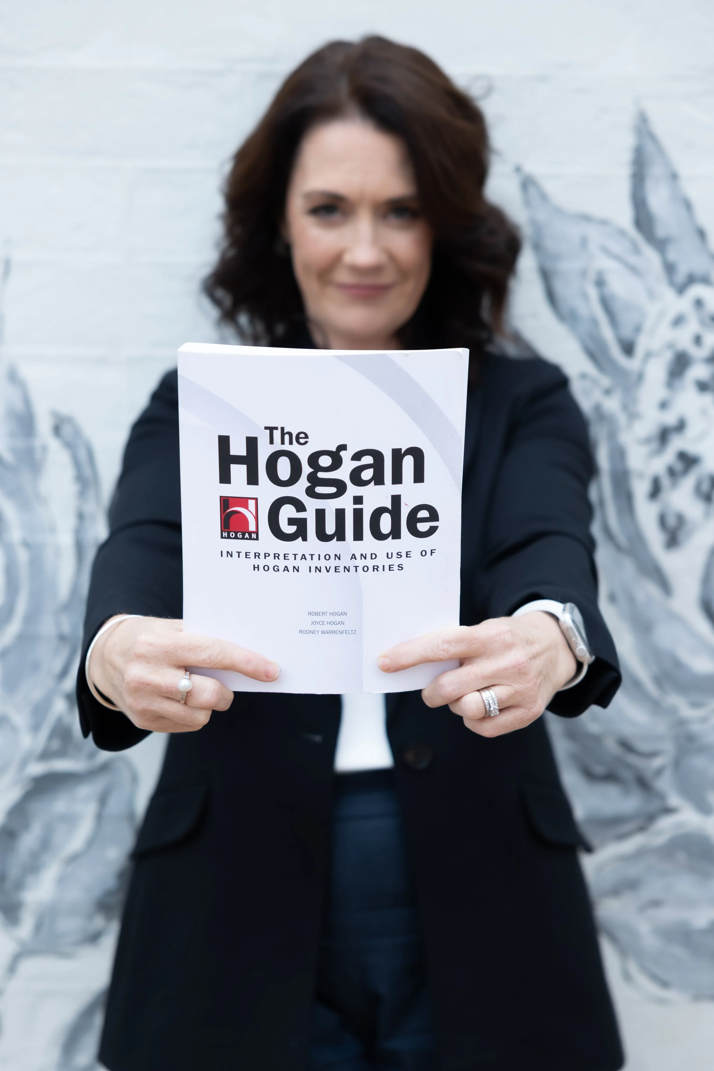 Louise Gibson · With Verve · executive leadership coach holding a book titled "The Hogan Guide" in front of her face, with a white wall featuring a painted eagle in the background.