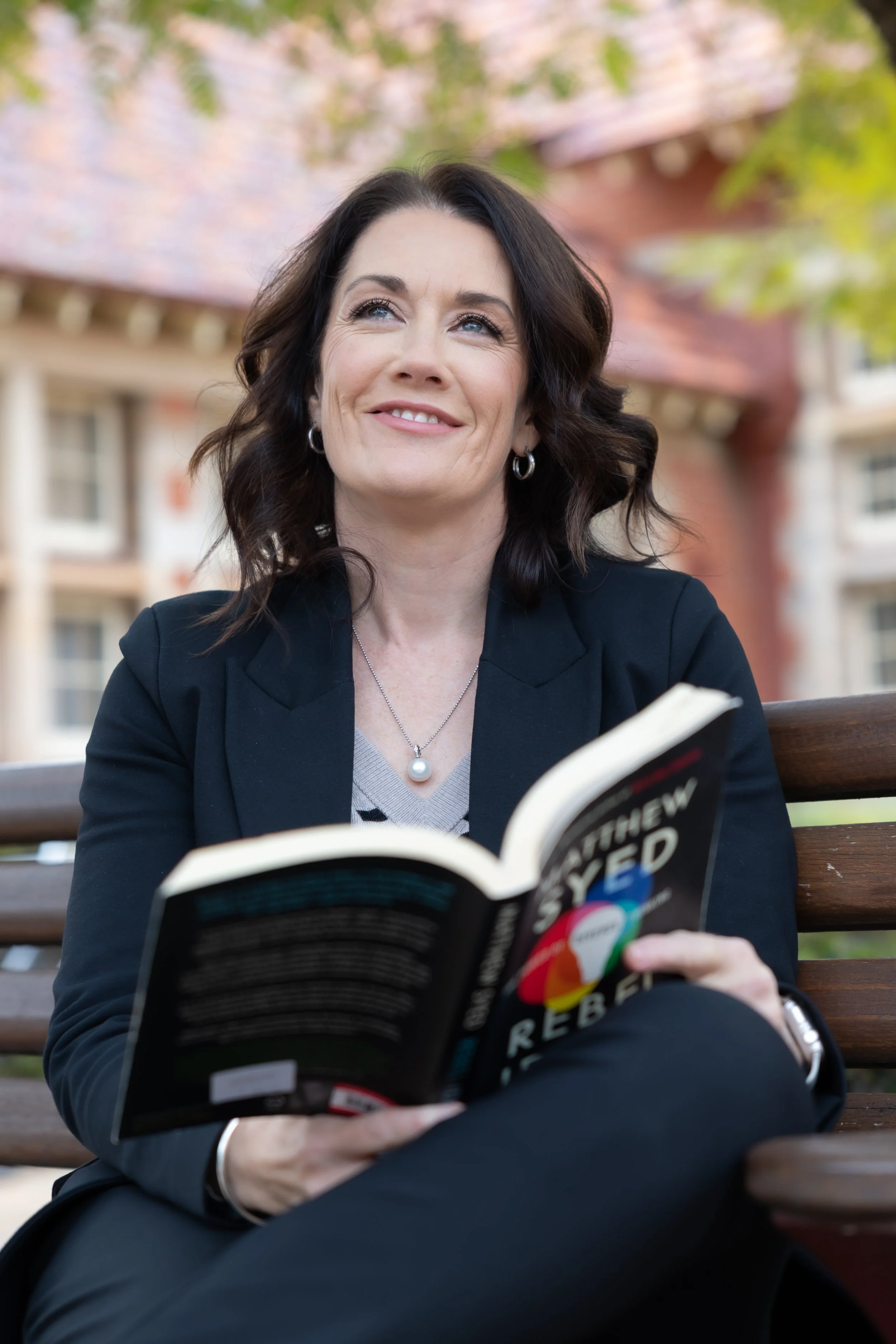 A woman with dark hair and earrings sitting on a park bench, smiling and reading a book titled 'Rebel'. She is wearing a dark blazer and a pearl necklace, with blurred trees and buildings in the background.