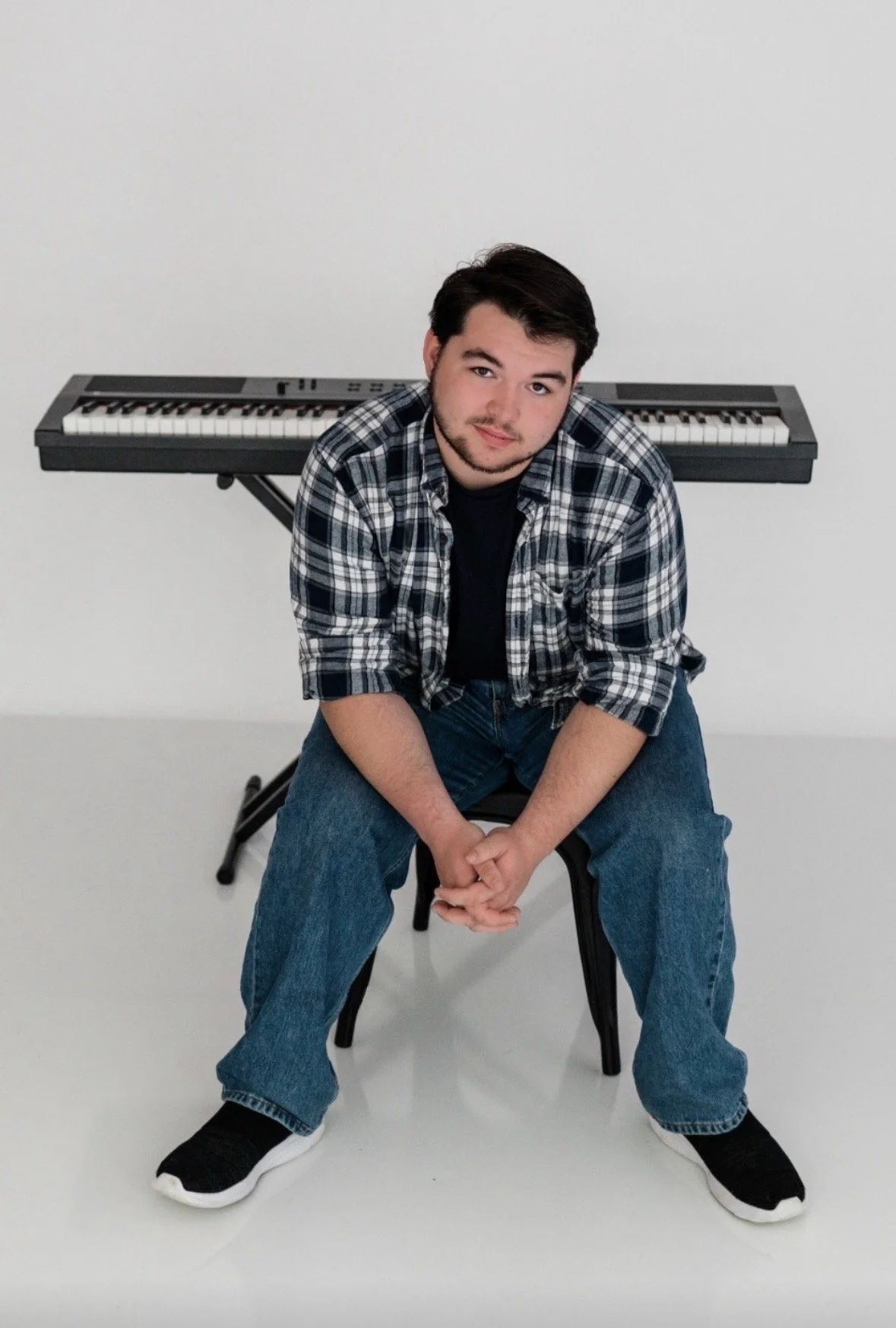 Image description of Jonathan: young man with dark hair and facial hair sitting on a chair in front of a keyboard. He's wearing a black T-shirt, plaid shirt overshirt, and jeans, with black sneakers, posing with an expression of confidence and poise.