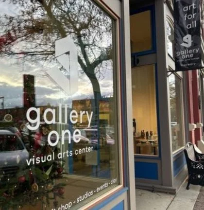 Storefront window with the logo of 'Gallery One Visual Arts Center' and a reflection of trees and cars outside.