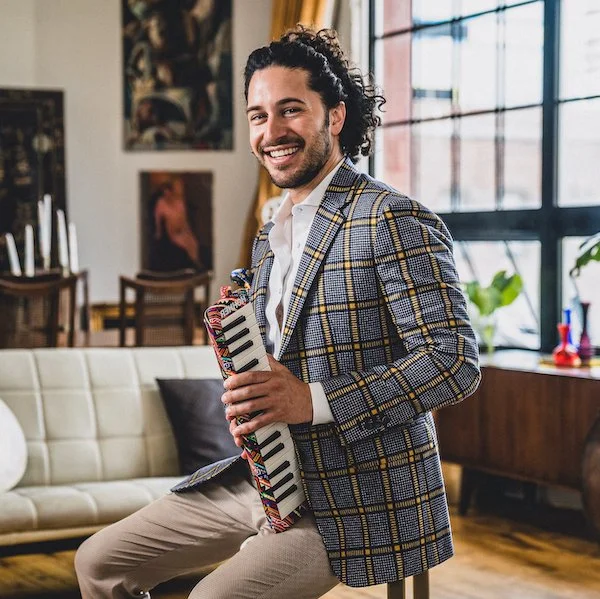 Image description of famous jazz pianist Emmet Cohen: A smiling man with dark curly hair and a beard, dressed in a plaid blazer and beige pants, sitting in a stylish living room while holding a colorful toy keyboard called a melodica.