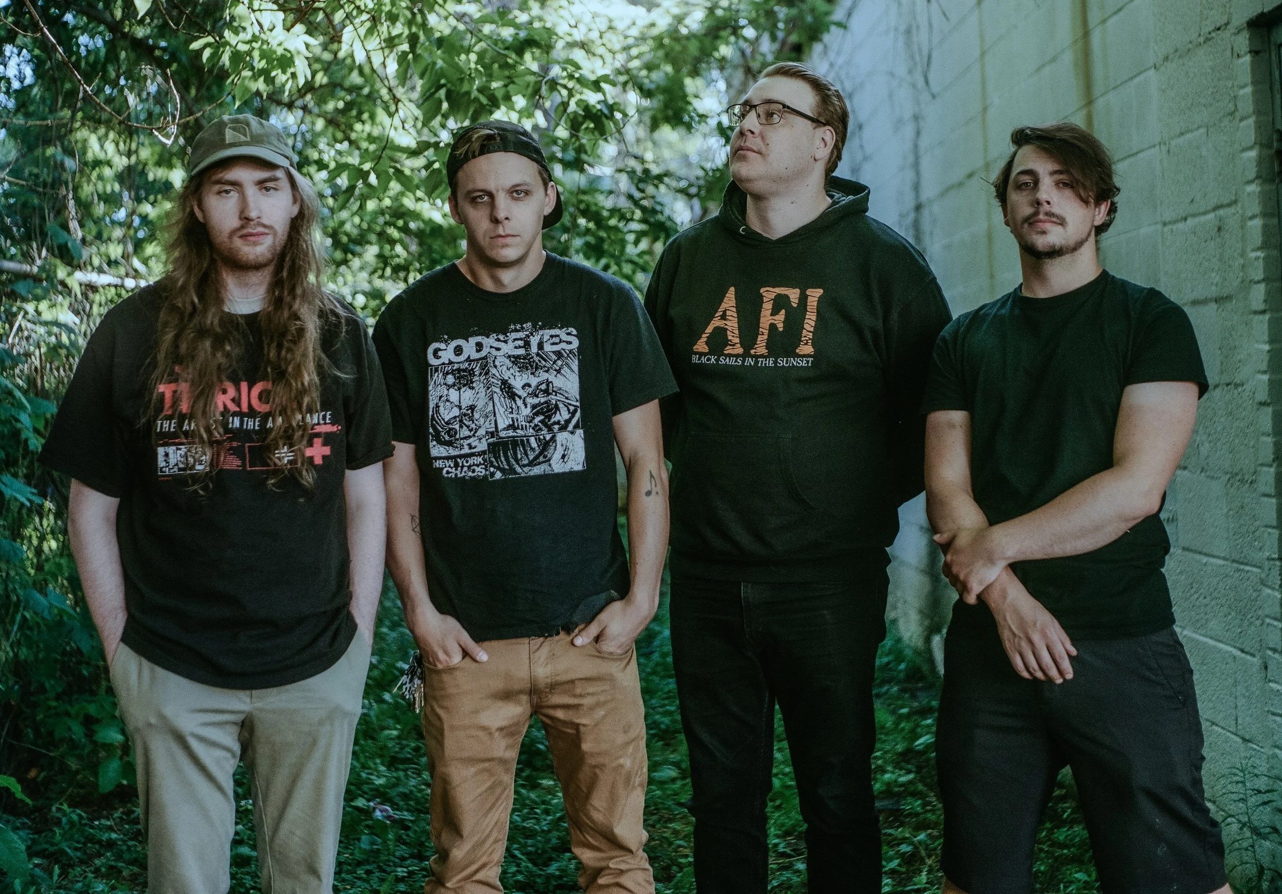 Four young men standing outdoors against a green leafy background and a gray wall, dressed casually with neutral expressions.