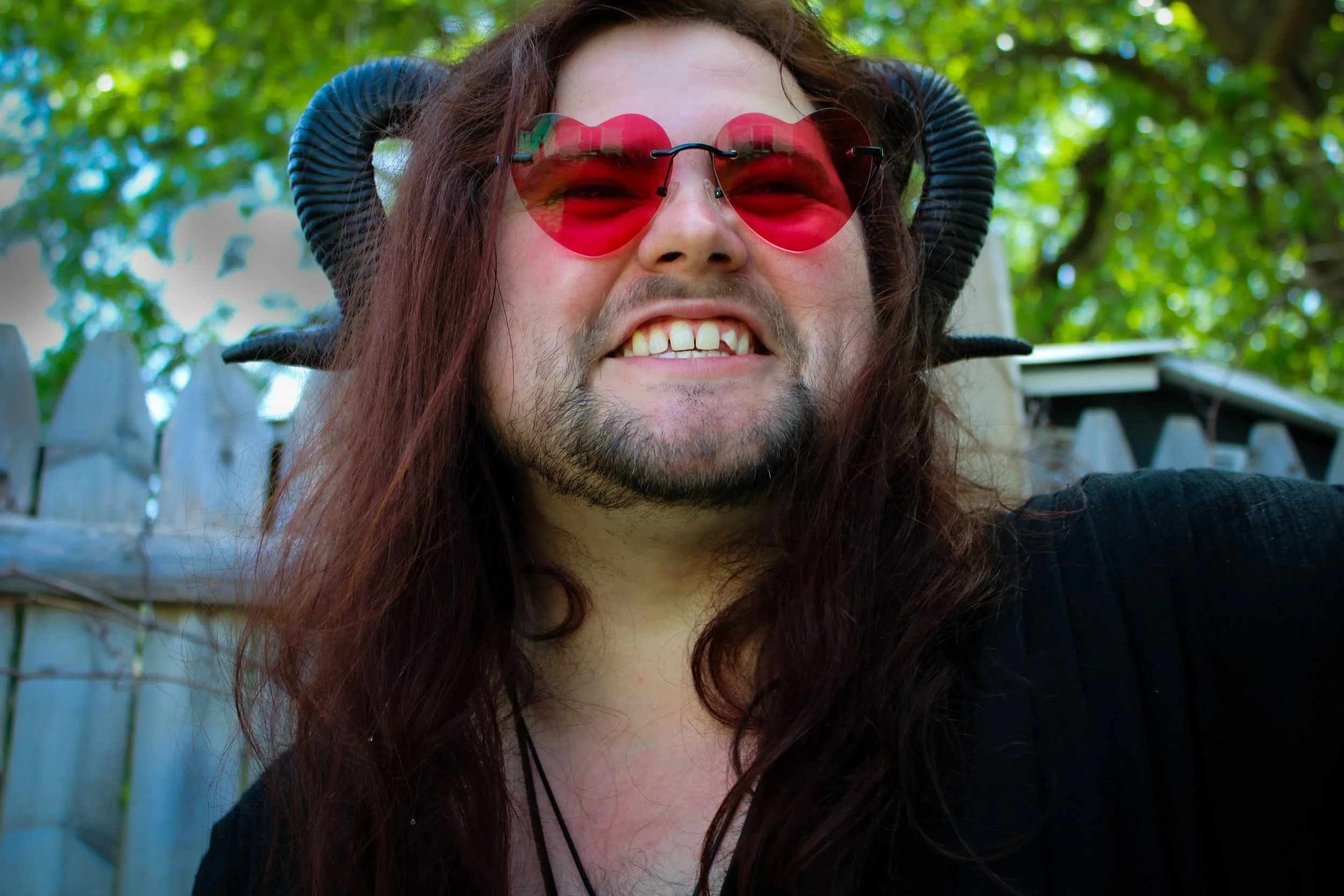 A person with long, wavy brown hair wearing heart-shaped red sunglasses, smiling with visible teeth, outdoors with green trees and a wooden fence in the background.
