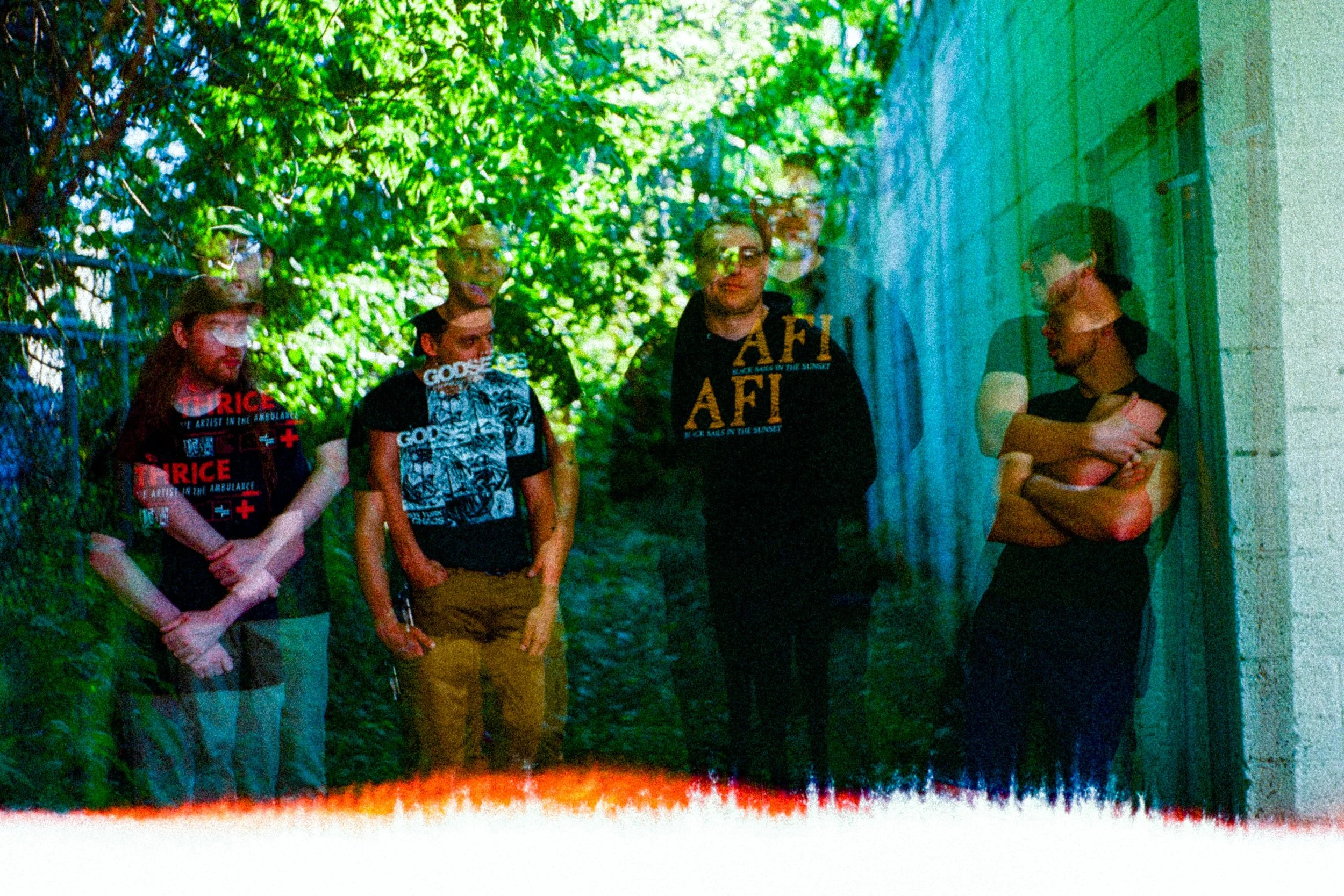 Five young people standing outdoors beneath green trees, with some wearing casual clothing and glasses, looking towards the camera. One person is leaning against a wall with arms crossed.