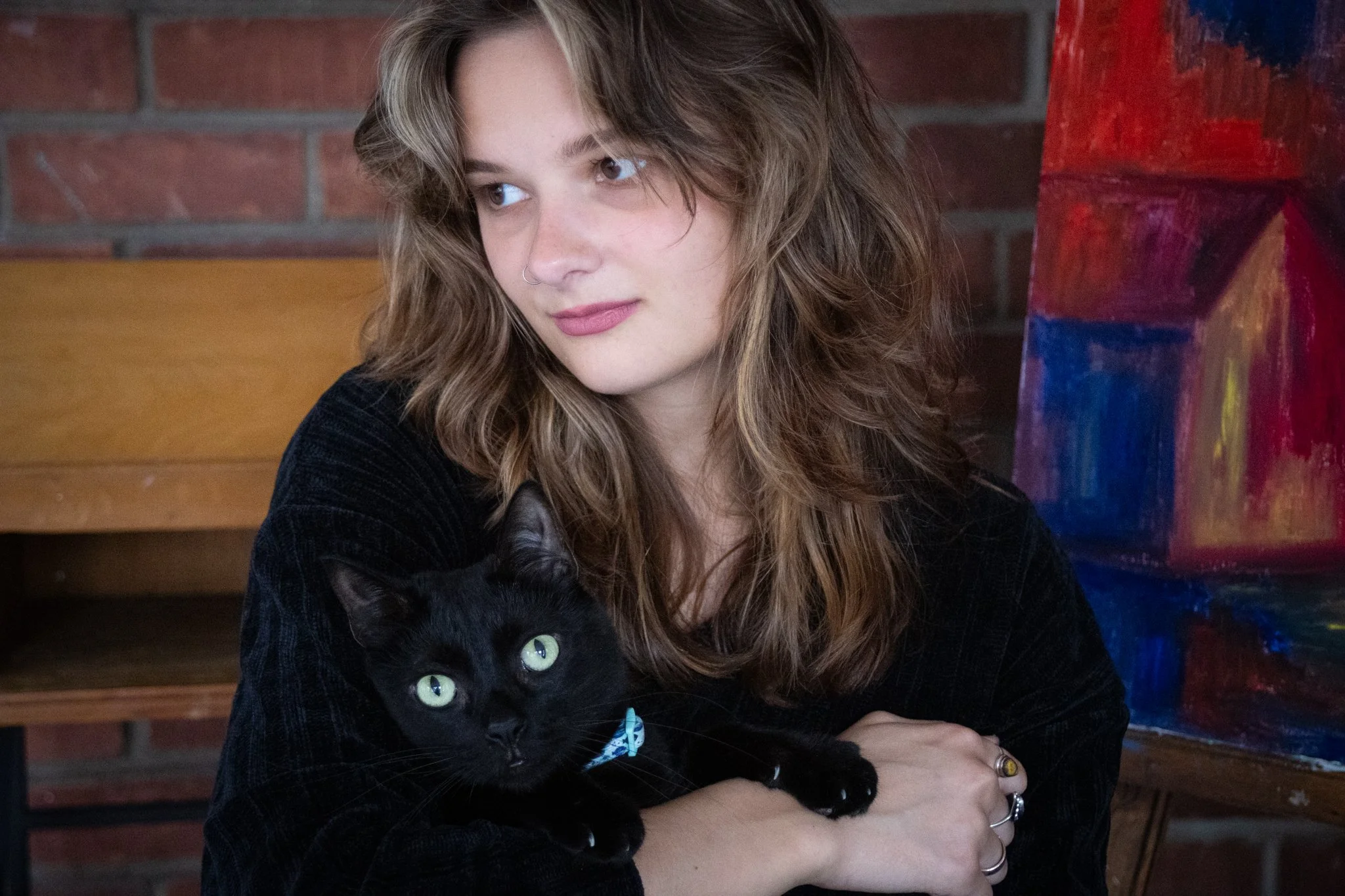 A young woman with wavy light brown hair holds a black cat with green eyes while sitting indoors against a brick wall and colorful artwork.